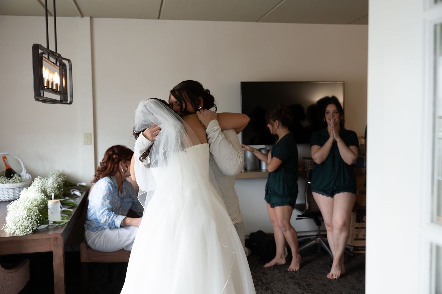 Bride in white ball gown having veil adjusted by bridesmaid while others react emotionally nearby.