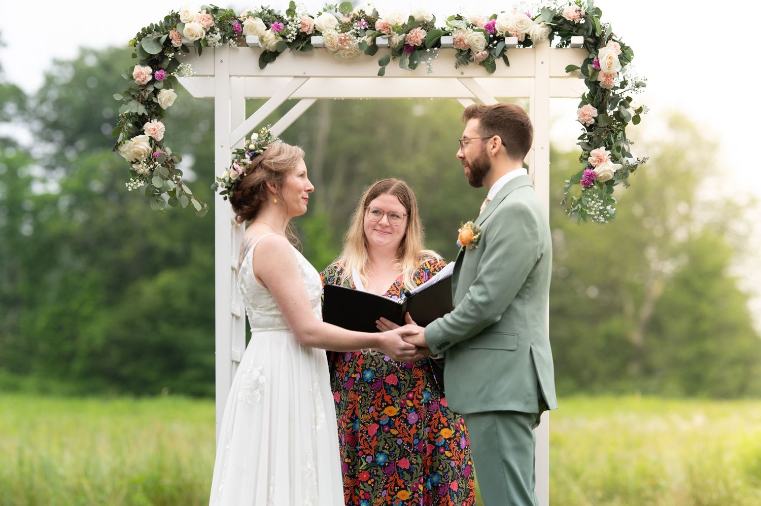 Bride and groom hold hands during outdoor wedding ceremony under floral arch with officiant.