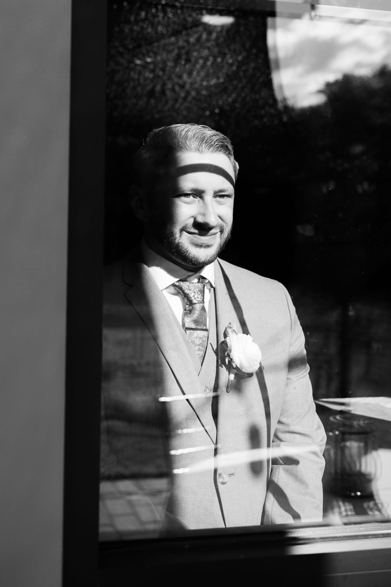 Black and white photo of a smiling groom in a suit with boutonniere, photographed through a window.