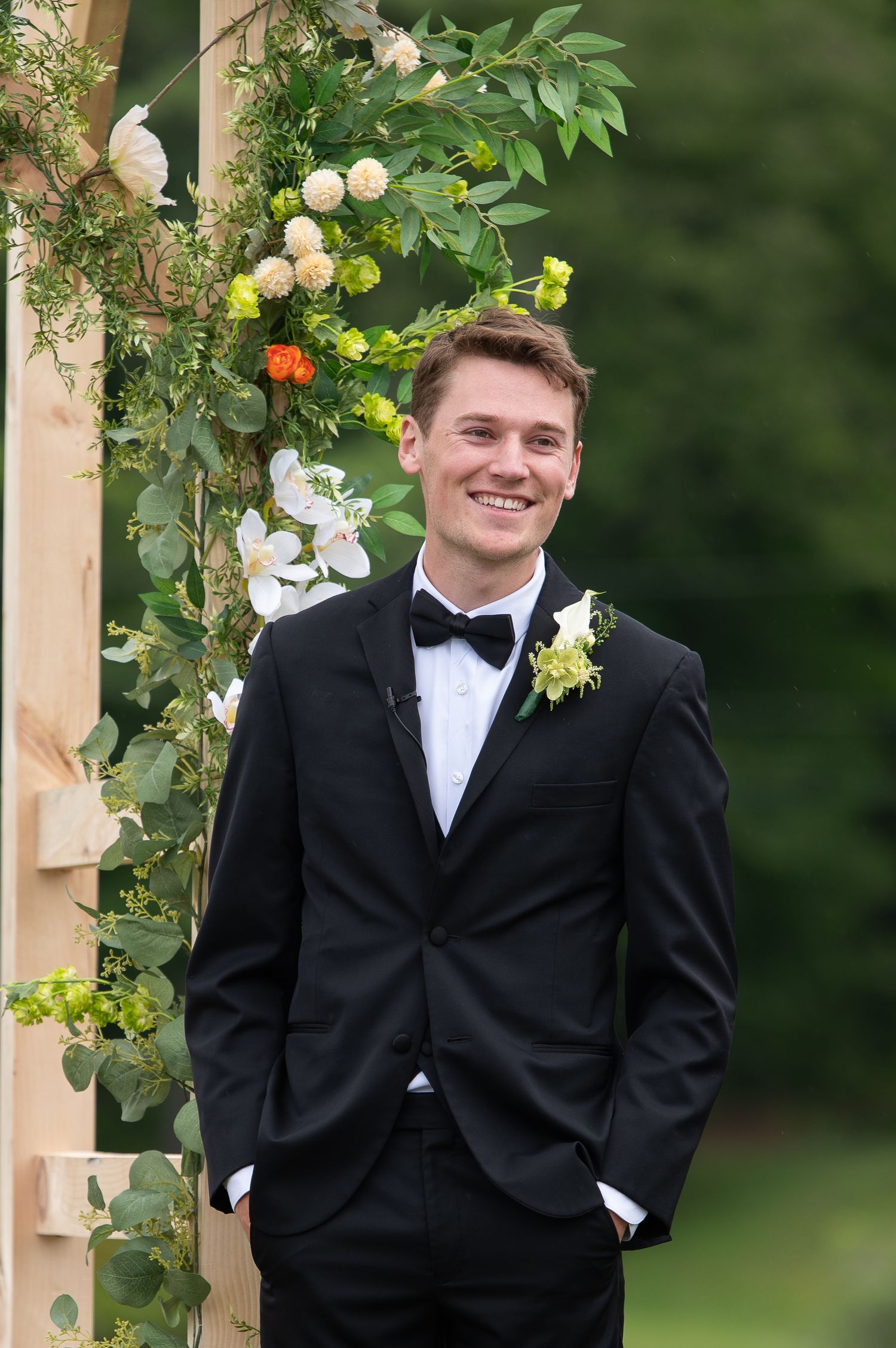 Smiling groom in black tuxedo with boutonniere stands before floral arch with greenery at outdoor wedding.