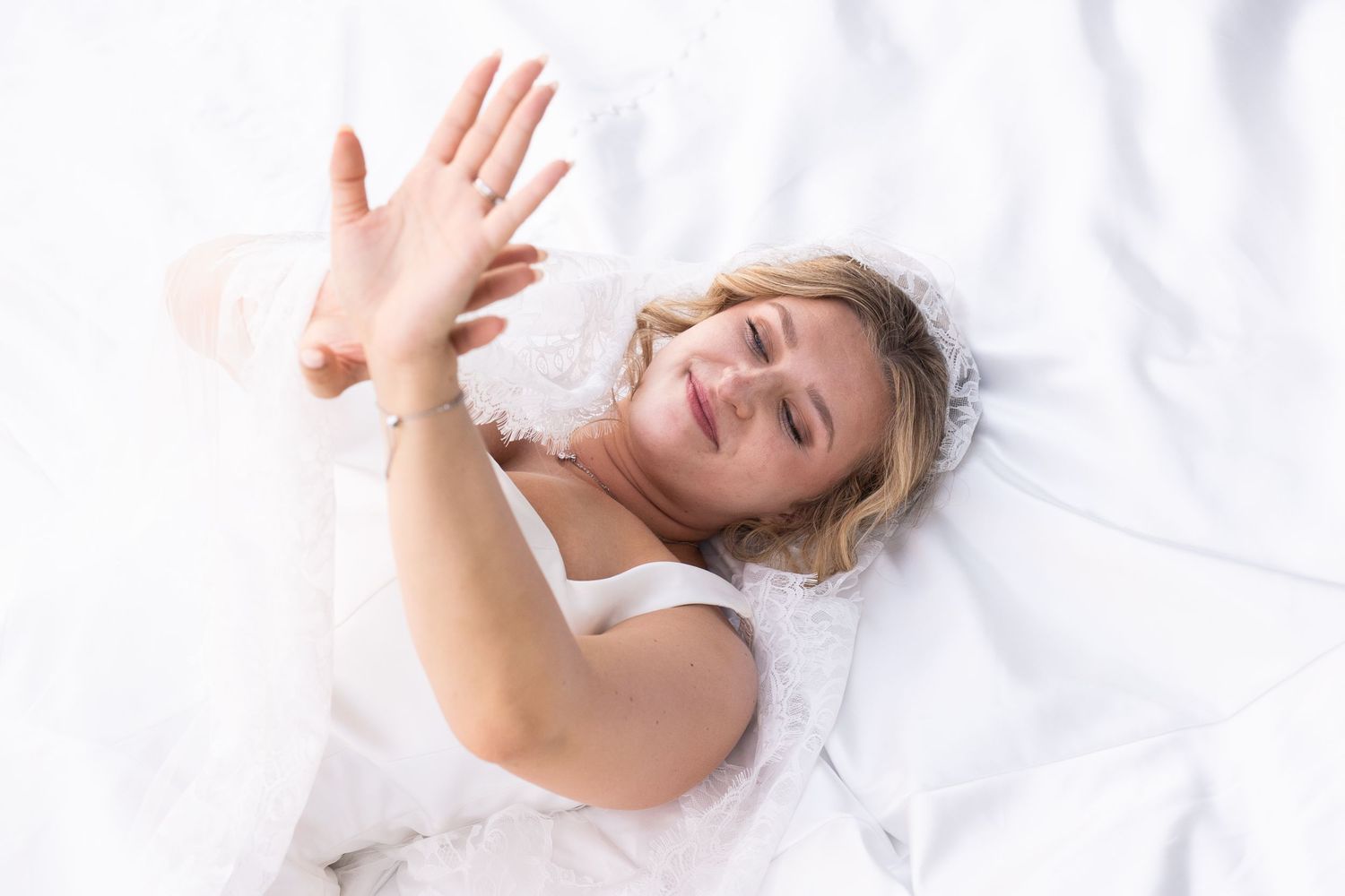 Woman lying on white bedding, raising her hand toward camera with a relaxed smile.