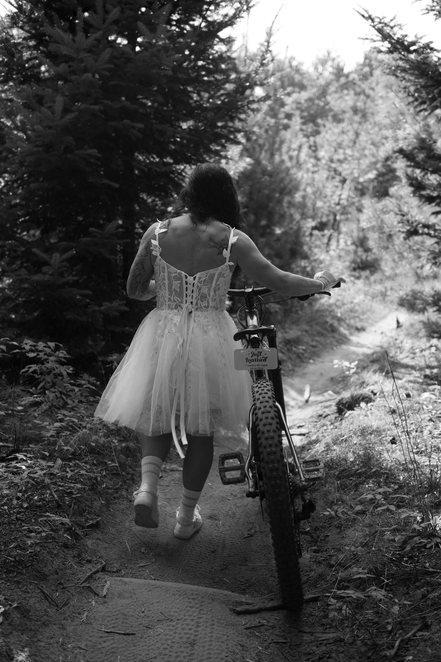Bride in white tulle dress and ballet flats walks mountain bike along forest trail in black and white photo.