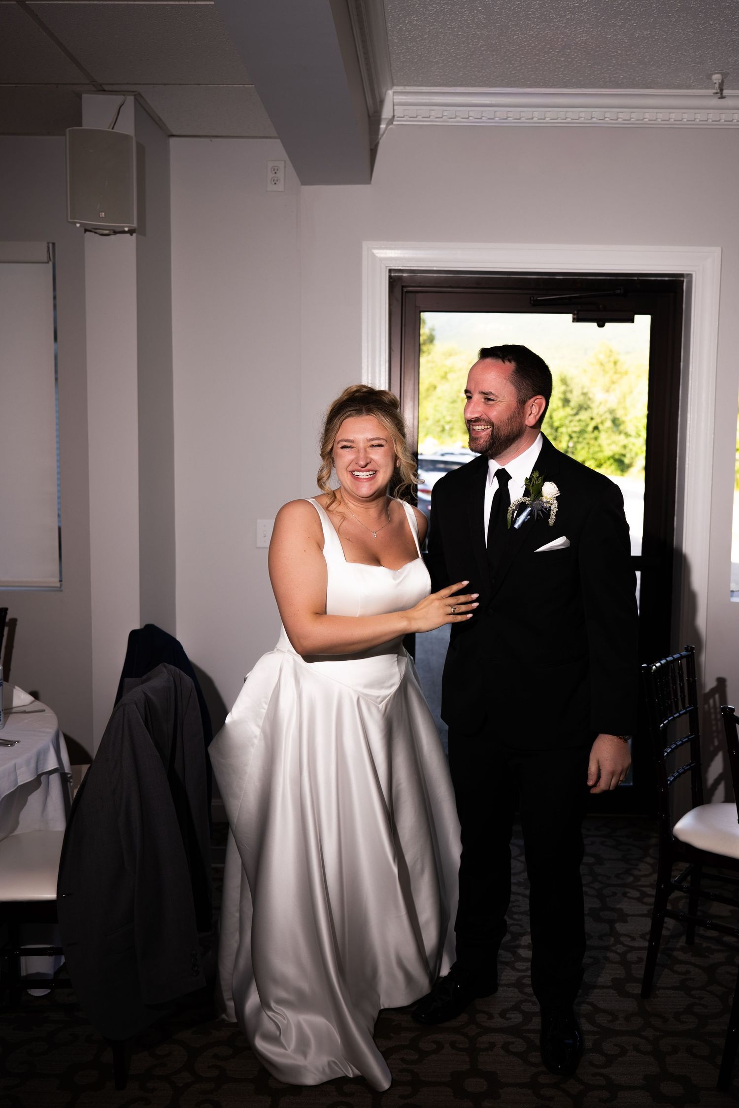 Bride in white gown and groom in black tuxedo smiling together indoors at their wedding reception.