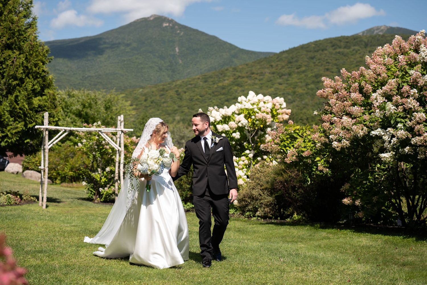 Bride and groom walk hand in hand on lush green lawn with mountain backdrop and blooming flowers on sunny wedding day.