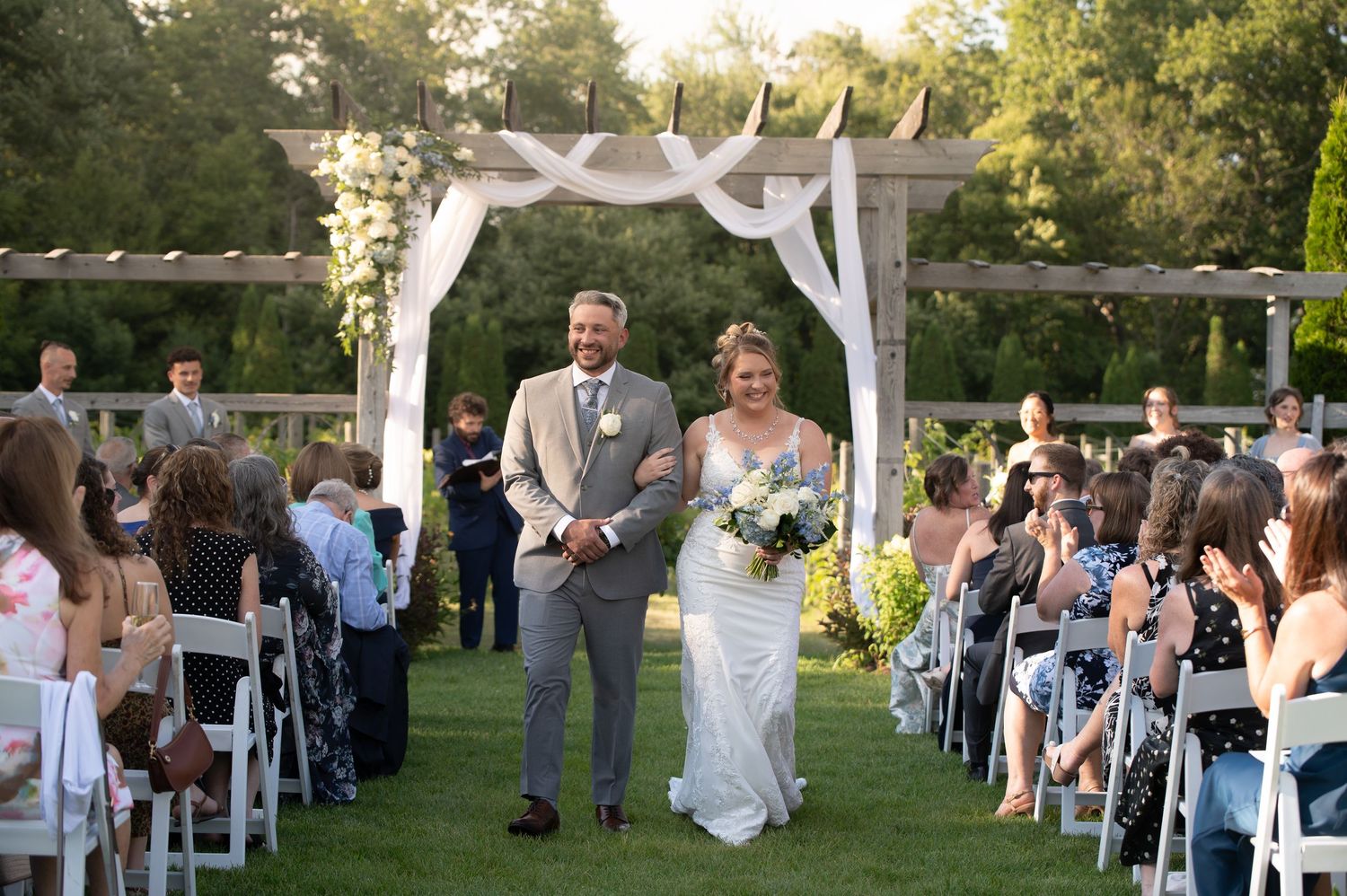 Newlywed couple walks down outdoor wedding aisle under white floral arch, surrounded by seated guests on a sunny day.