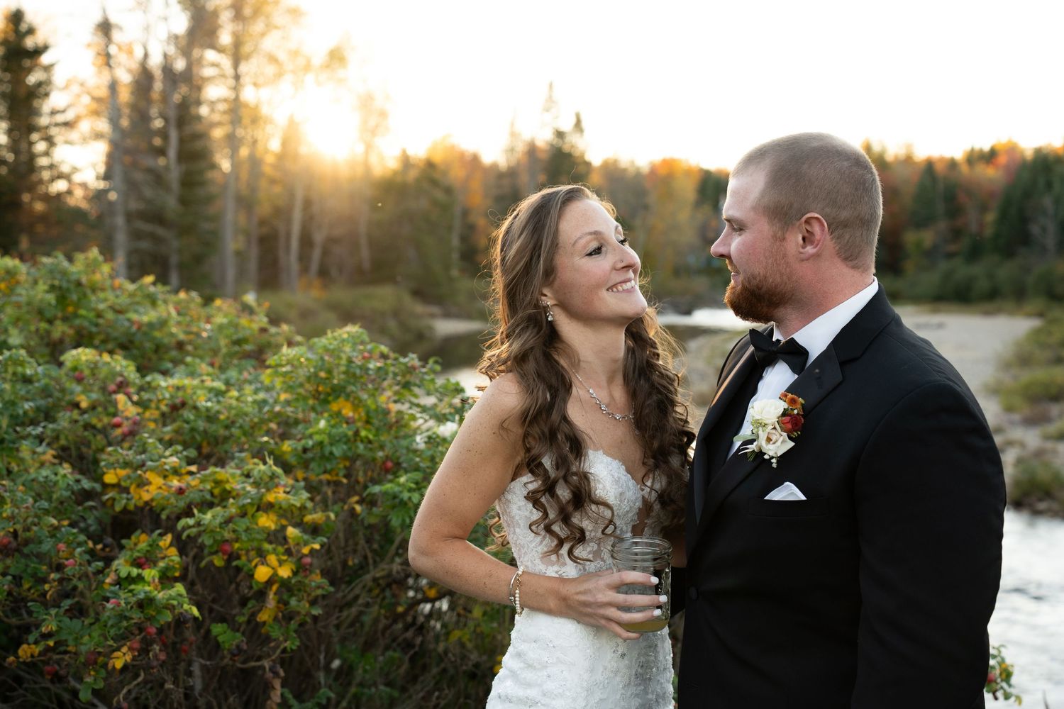 Bride and groom share a romantic moment at sunset near a river surrounded by autumn foliage in a forest setting.