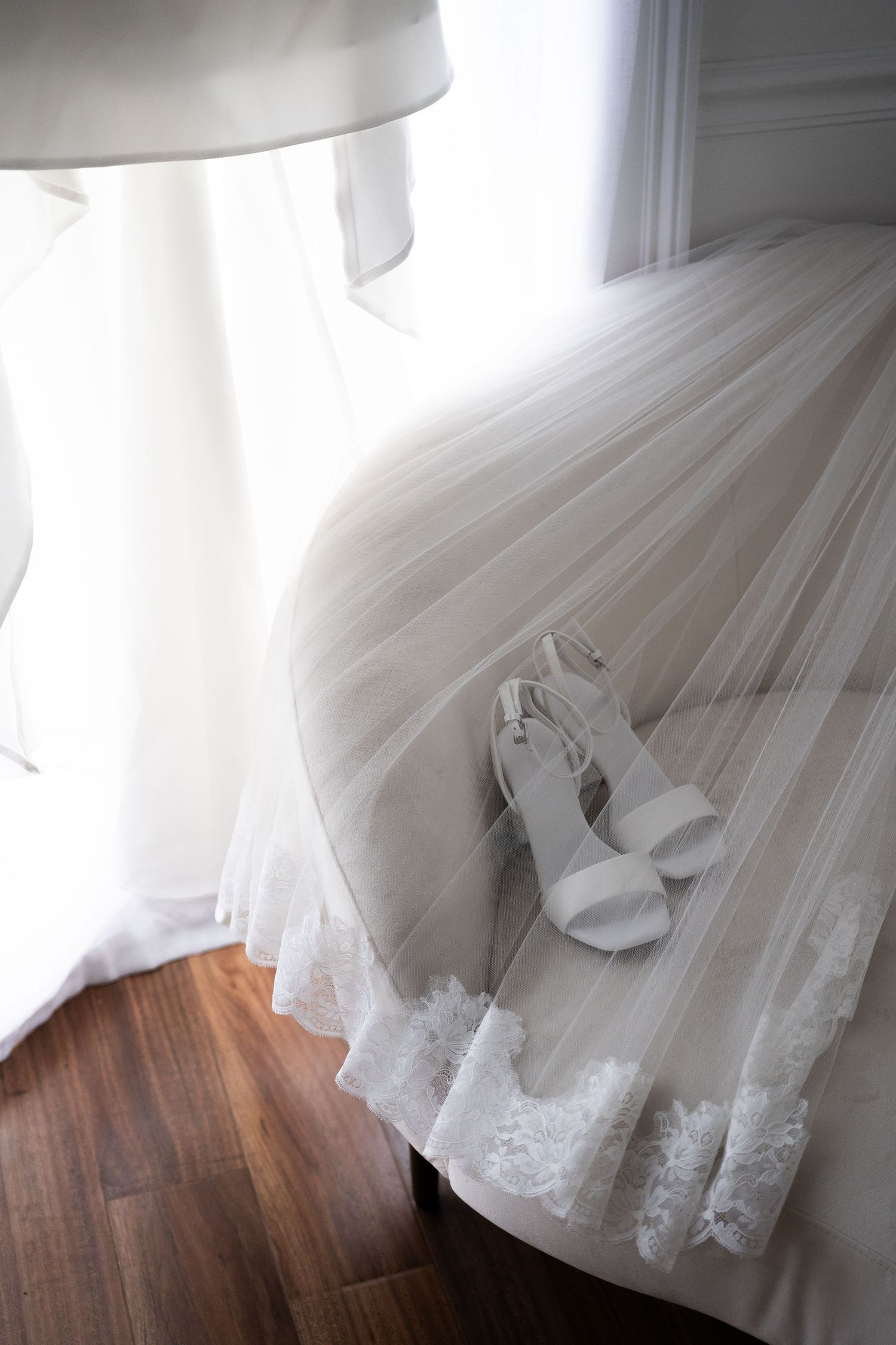 White wedding dress with lace hem and bridal heels displayed on chair near window.