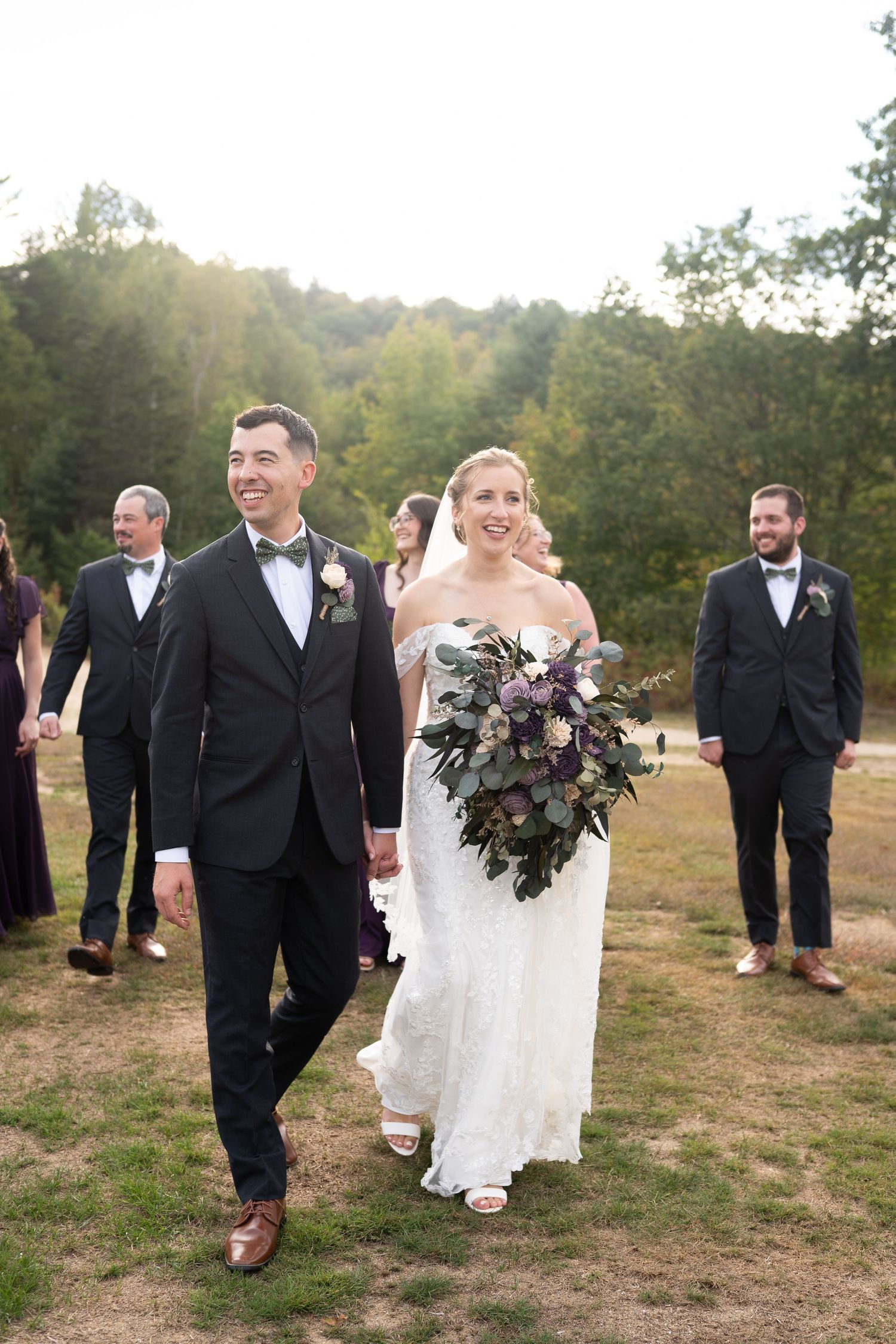 Smiling bride and groom walking with wedding party in a sunny outdoor field after ceremony.