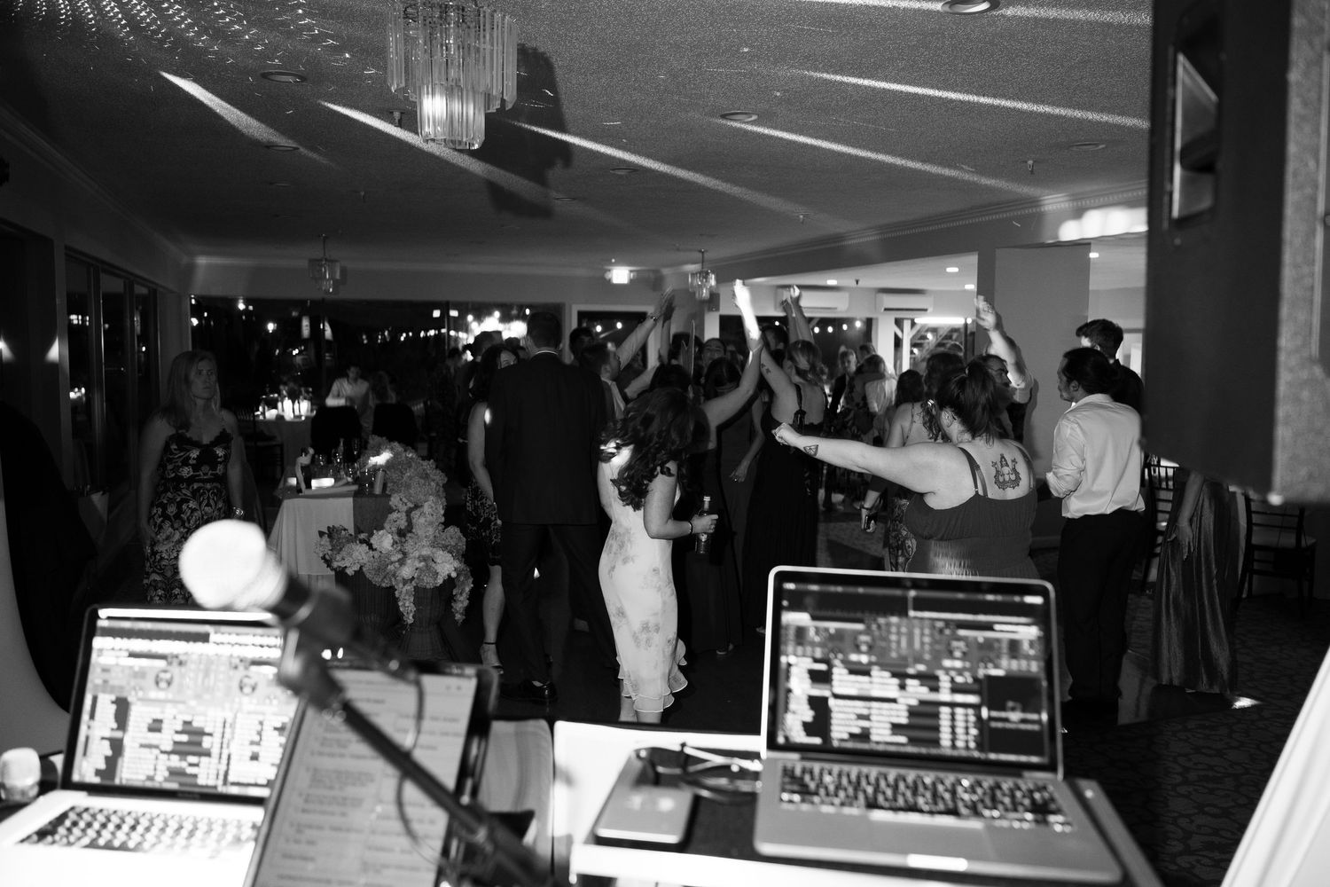Black and white photo of a lively DJ setup with guests dancing at an indoor party or wedding reception.