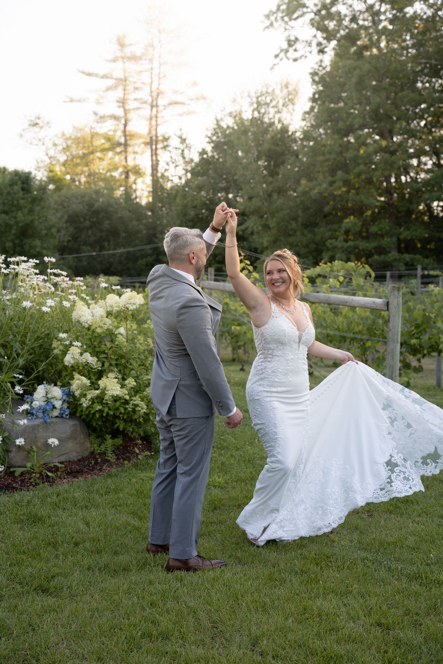Bride and groom dancing outdoors in a lush garden at sunset, bride in lace gown, groom in gray suit.