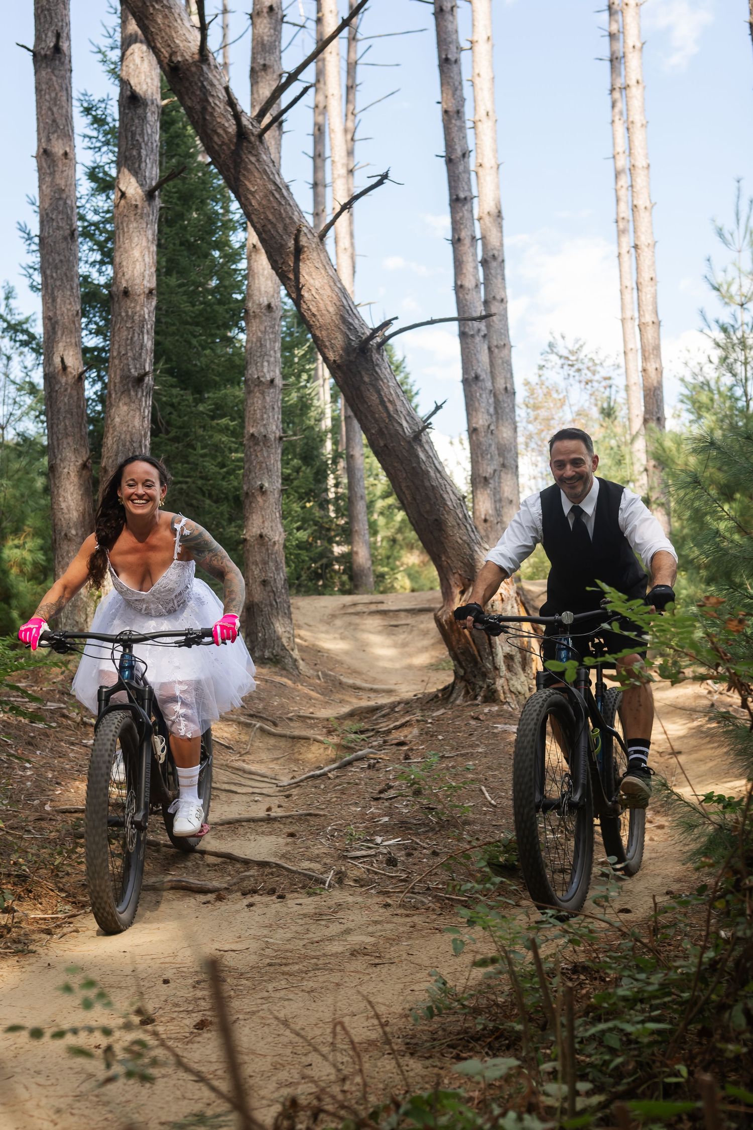 Laughing bride and groom in wedding attire ride mountain bikes side by side on a forest trail.