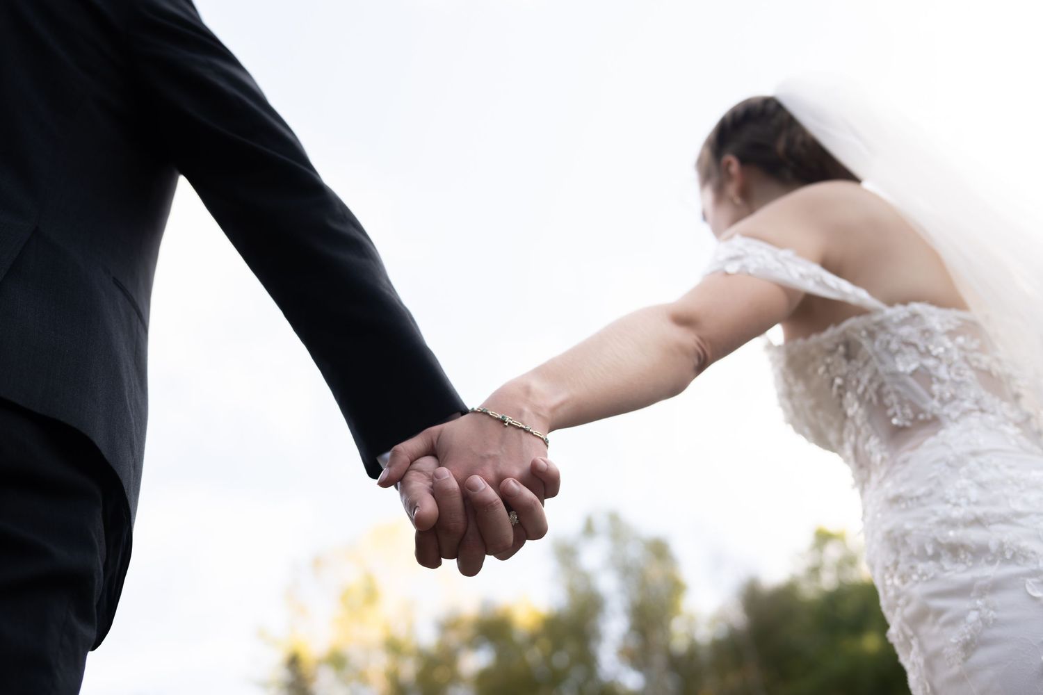 Bride and groom hold hands against bright sky in close-up shot showing lace wedding dress detail.
