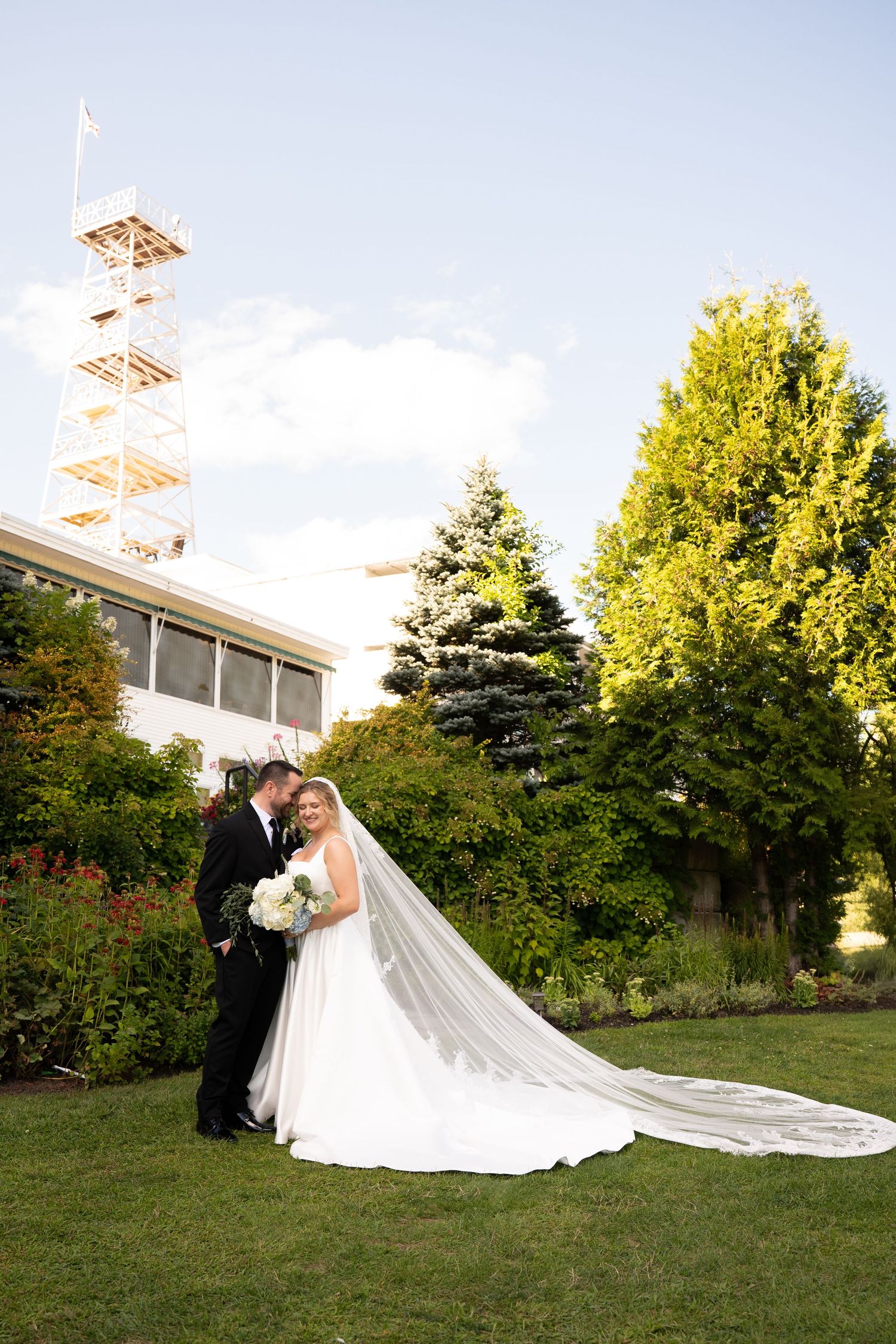 Bride and groom pose on green lawn with long veil trailing, trees and tower in background on sunny wedding day.