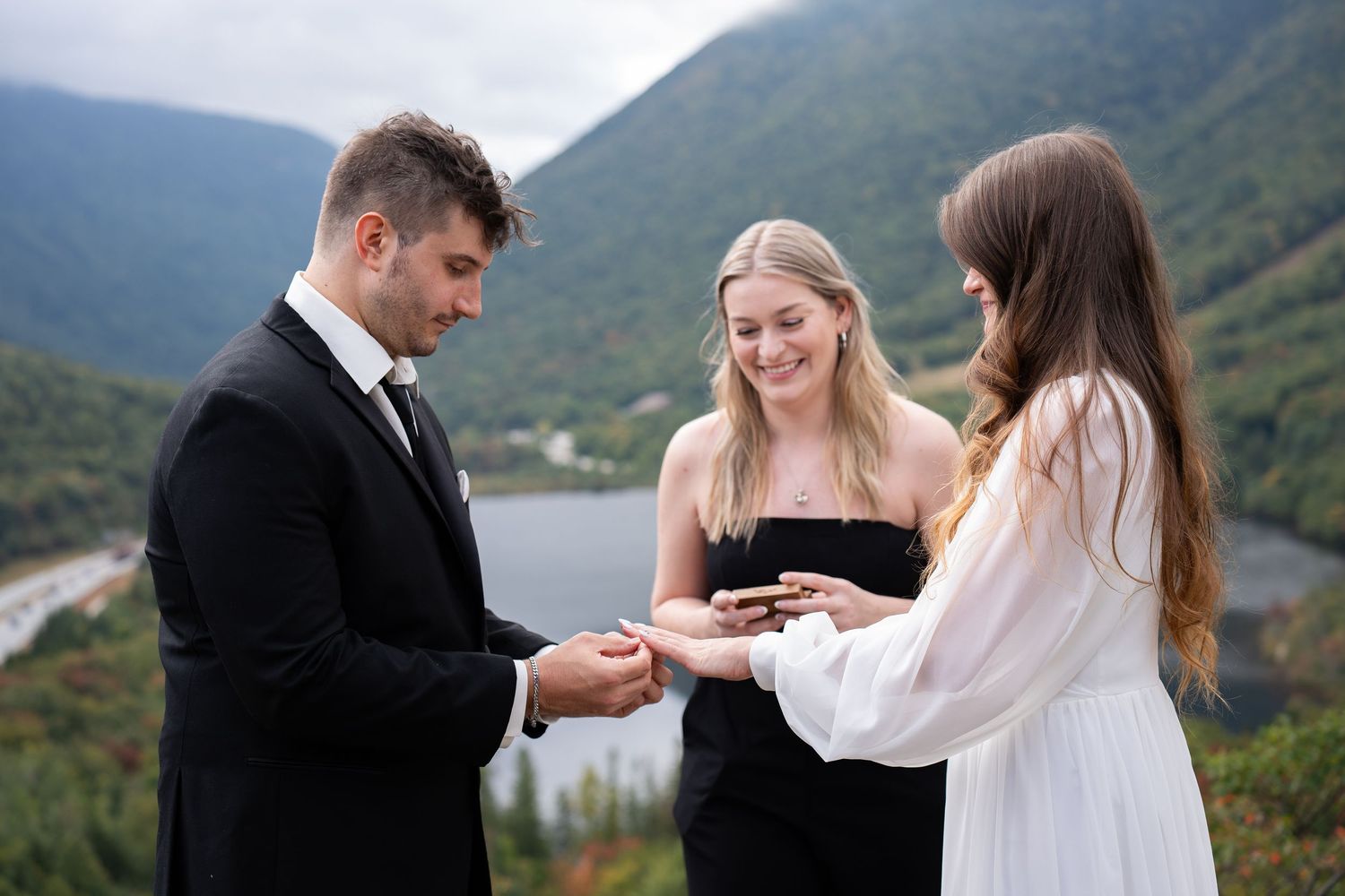 Couple exchanging rings during intimate mountain wedding ceremony with officiant overlooking scenic valley and lake.