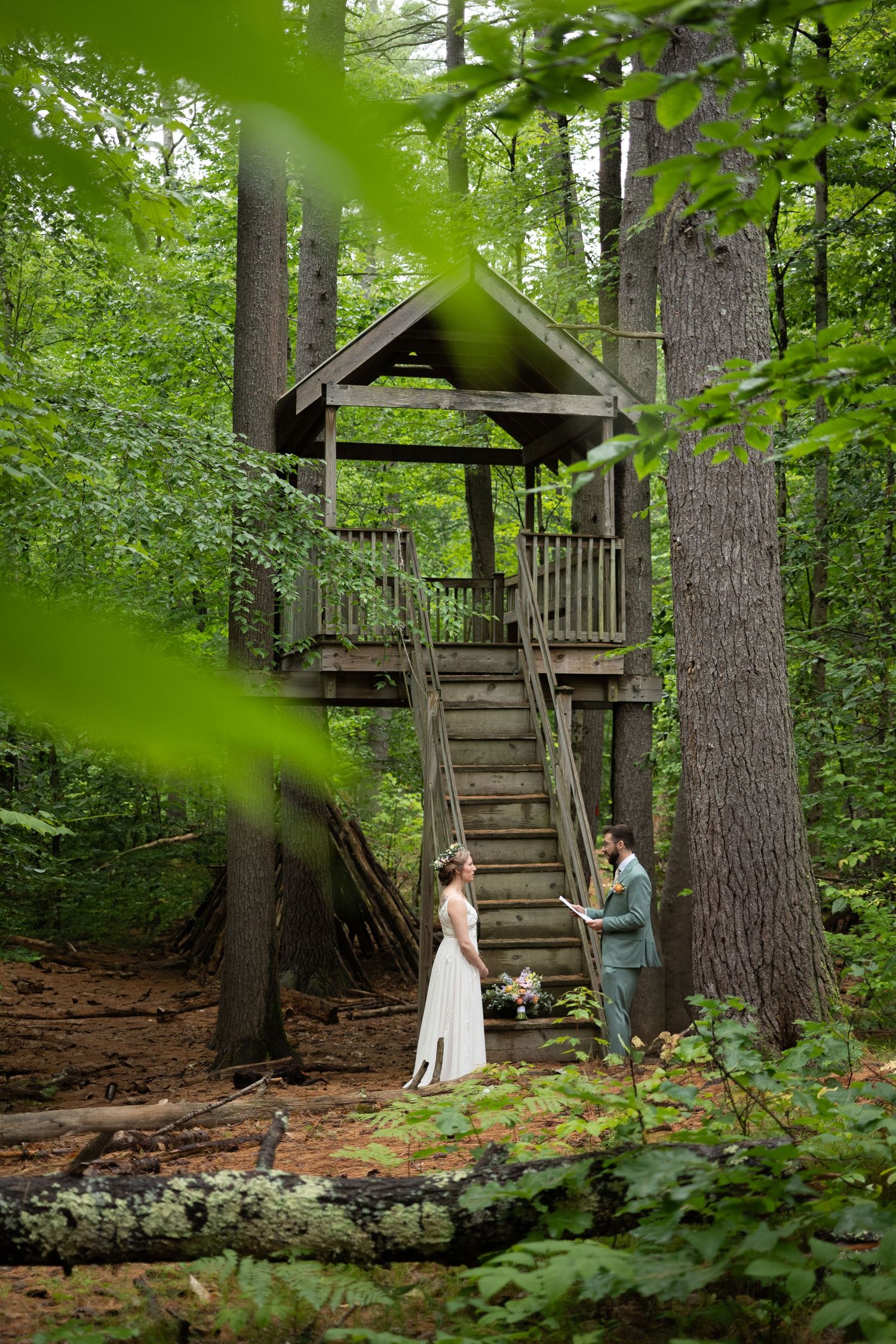 Bride and groom exchange vows beside a rustic wooden treehouse in a lush forest.