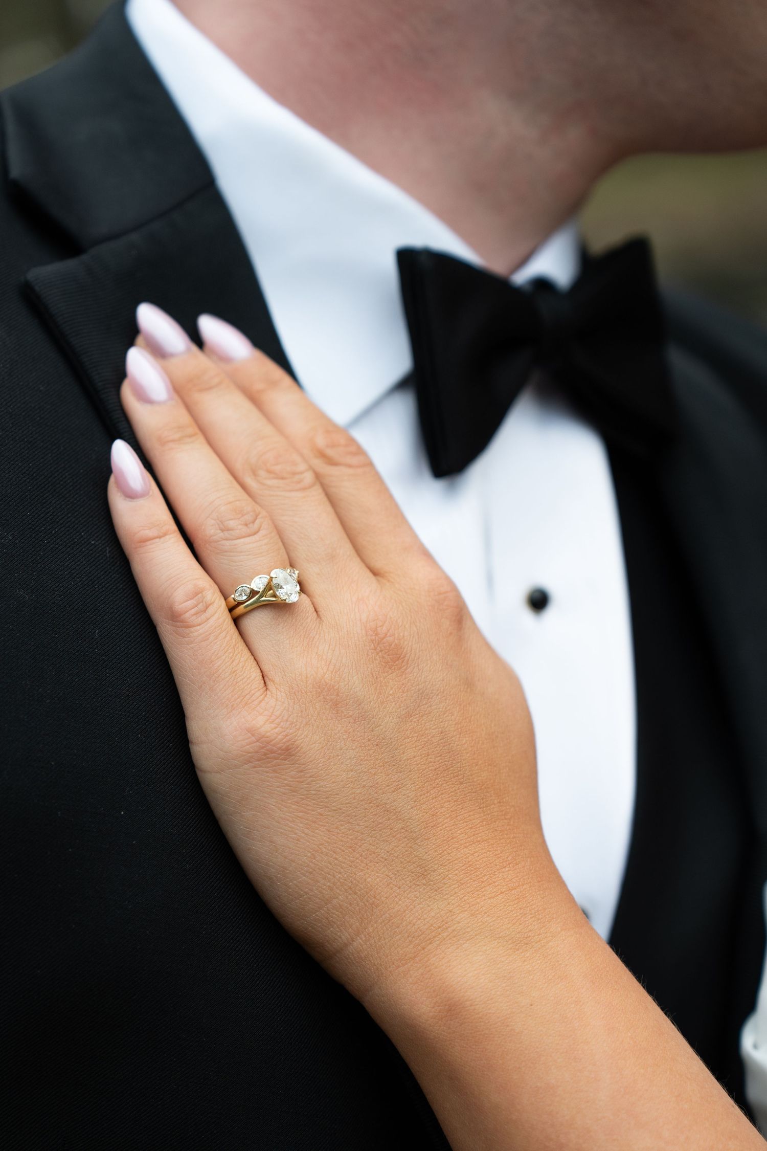 Close-up of bride's hand with gold diamond ring resting on groom's black tuxedo with bow tie.