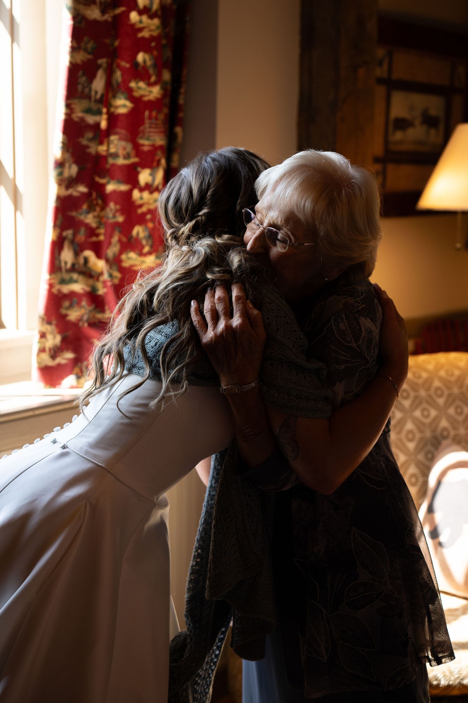 A bride in white lace gown shares a tender embrace with an elderly woman near a sunlit window indoors.