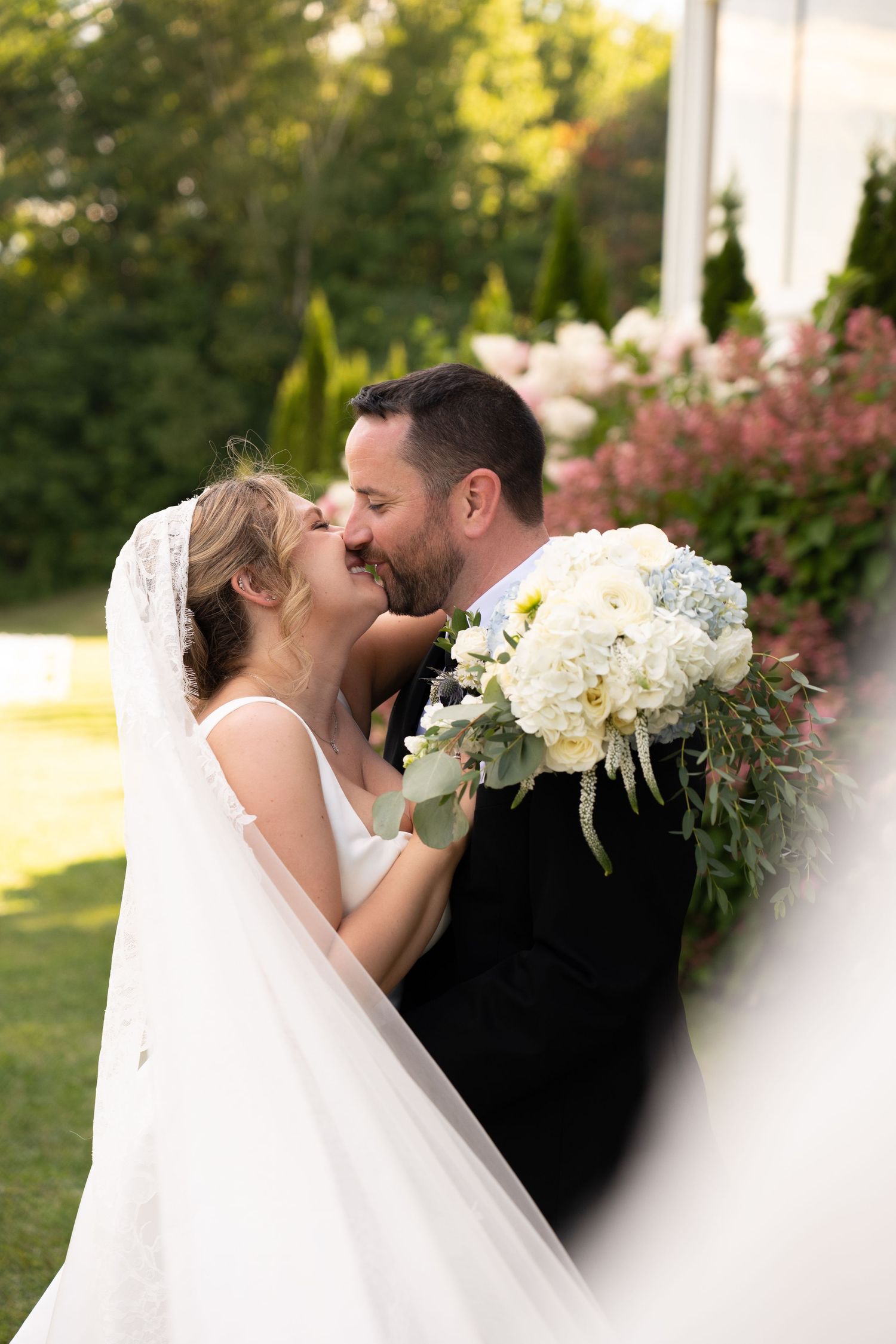 Bride and groom sharing a romantic kiss outdoors, bride holding white hydrangea bouquet with greenery on wedding day.