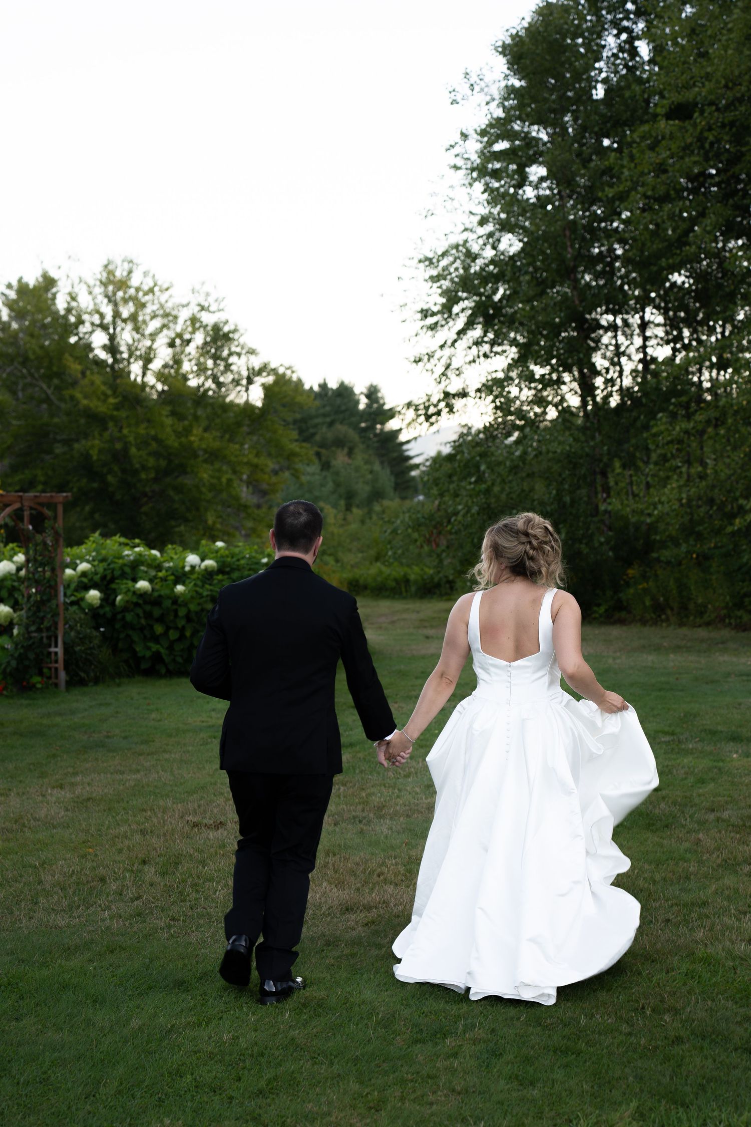 Bride and groom walking hand in hand across green lawn, viewed from behind, surrounded by lush trees and gardens.