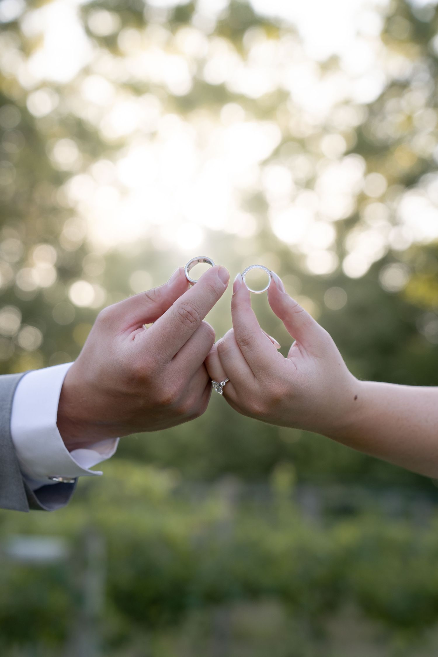 Bride and groom holding wedding rings up together outdoors with sunlit green trees bokeh background.