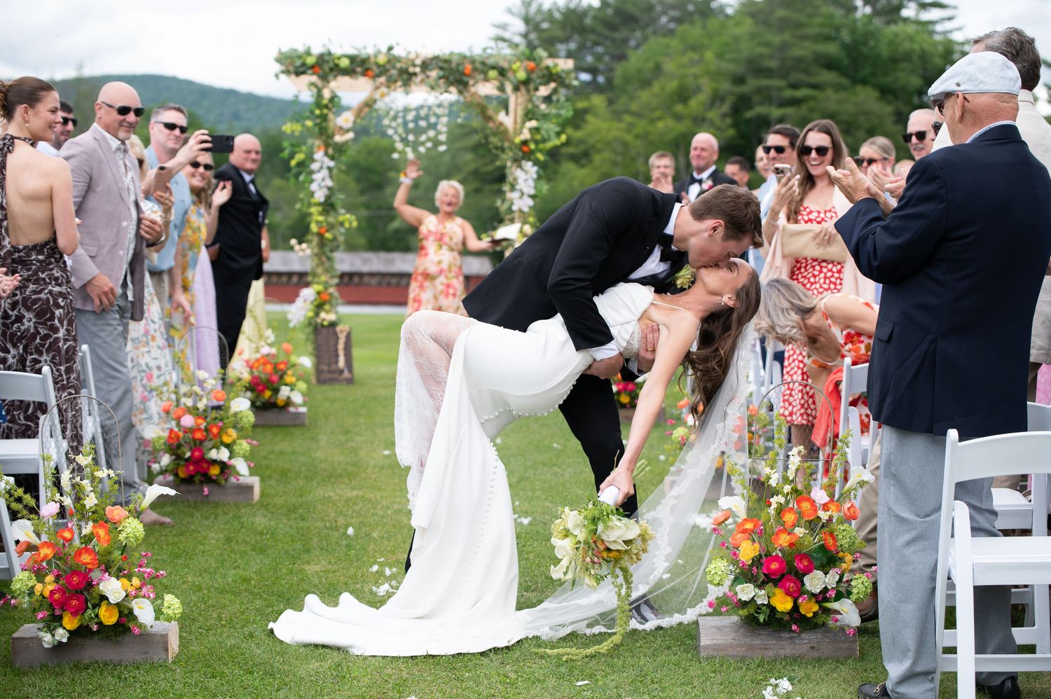 Groom dips bride for a romantic kiss at outdoor wedding ceremony with colorful floral arrangements and guests applauding.