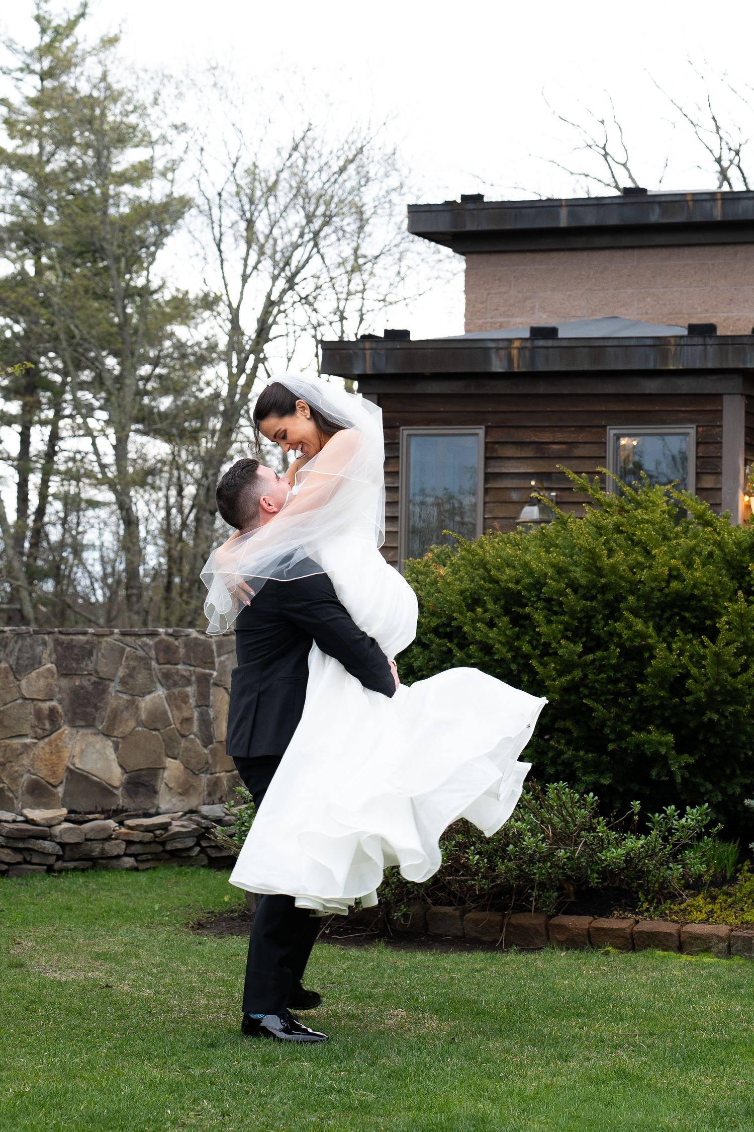 Groom in black tuxedo lifting bride in white ball gown and veil outdoors near rustic stone building.