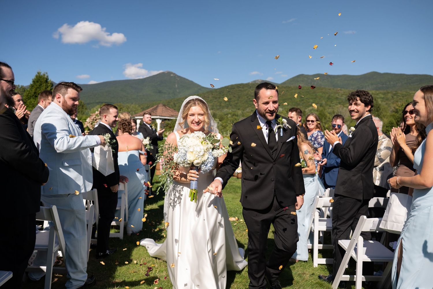 Bride and groom walk down outdoor aisle as guests toss flower petals, with mountain backdrop on sunny day.