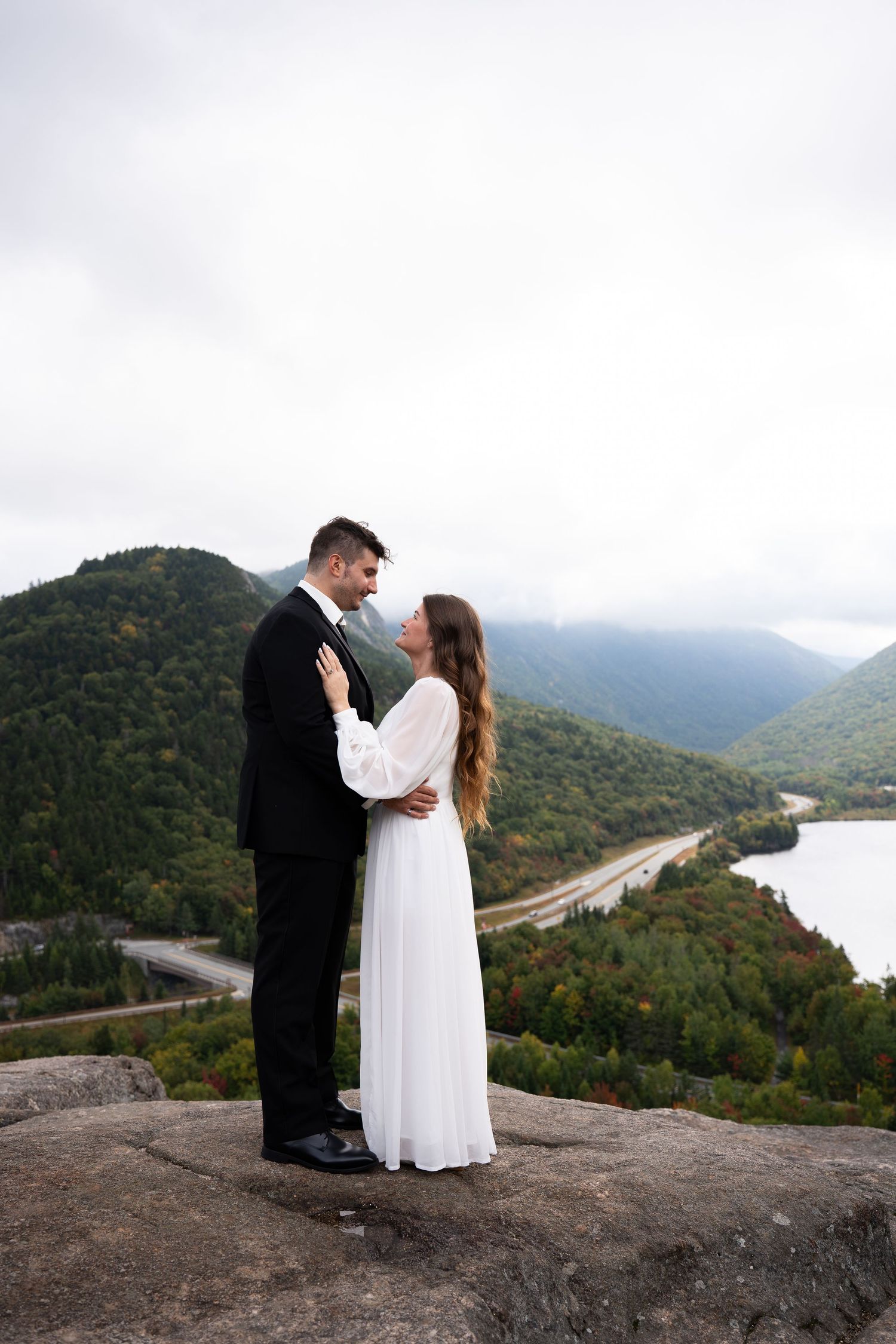 Bride and groom embrace on a mountain summit with scenic valley, river, and misty green hills in the background.