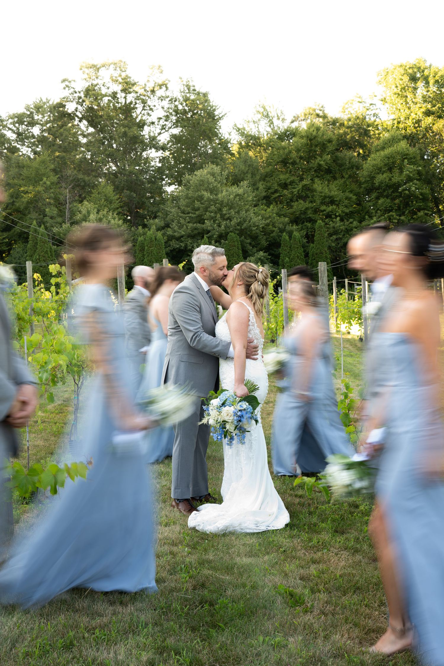 Bride and groom share a kiss surrounded by blurred bridesmaids in blue gowns at an outdoor vineyard wedding.