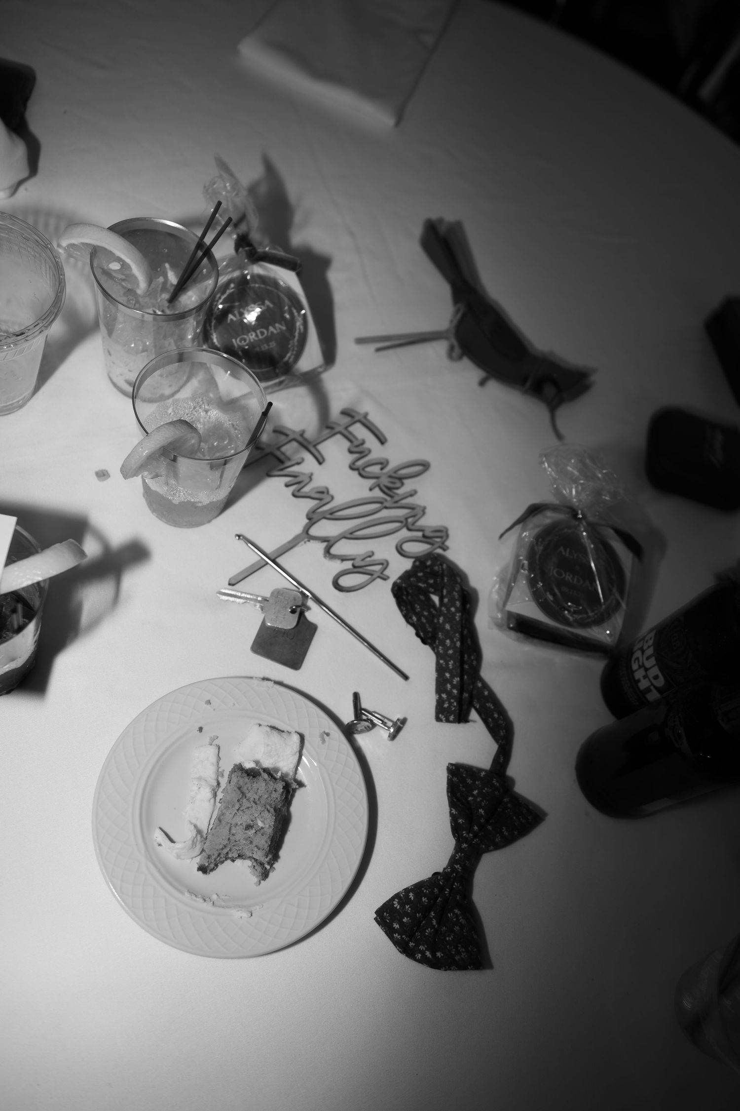 Black and white overhead shot of wedding table with cake, bow ties, glasses and decor remnants.