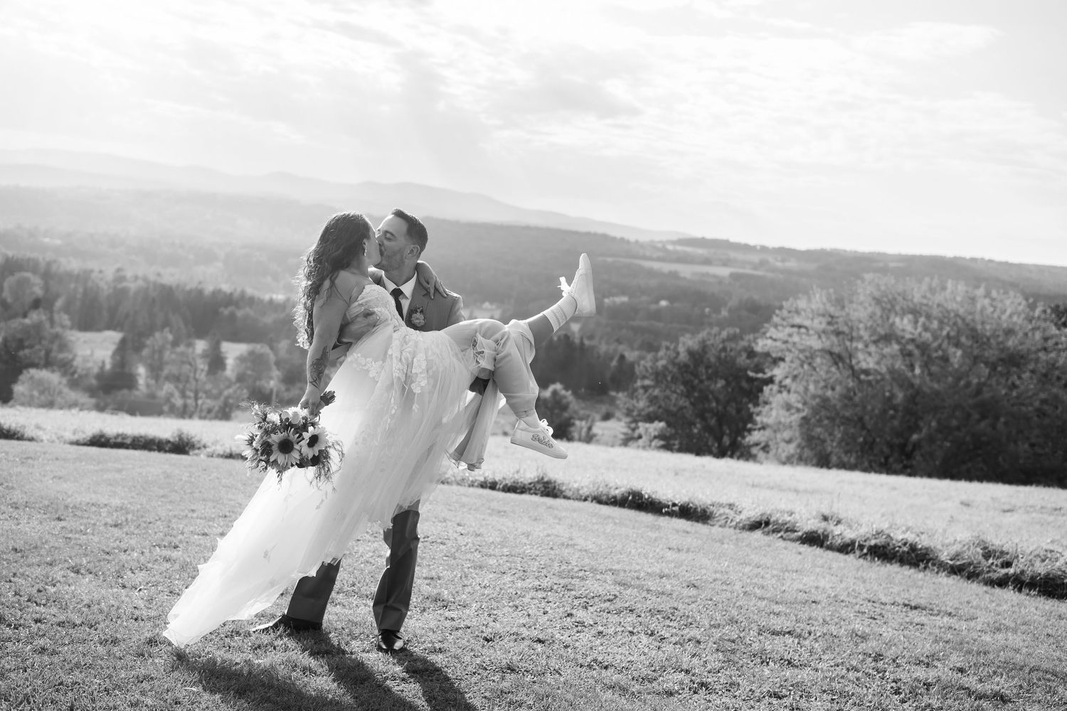 Black and white photo of groom lifting bride in a field with scenic countryside views.