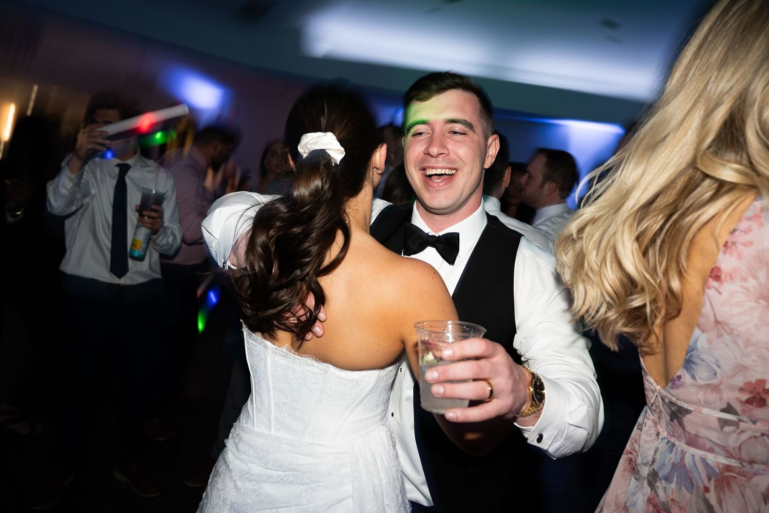 Bride and groom dance together joyfully with guests at colorfully lit wedding reception party.