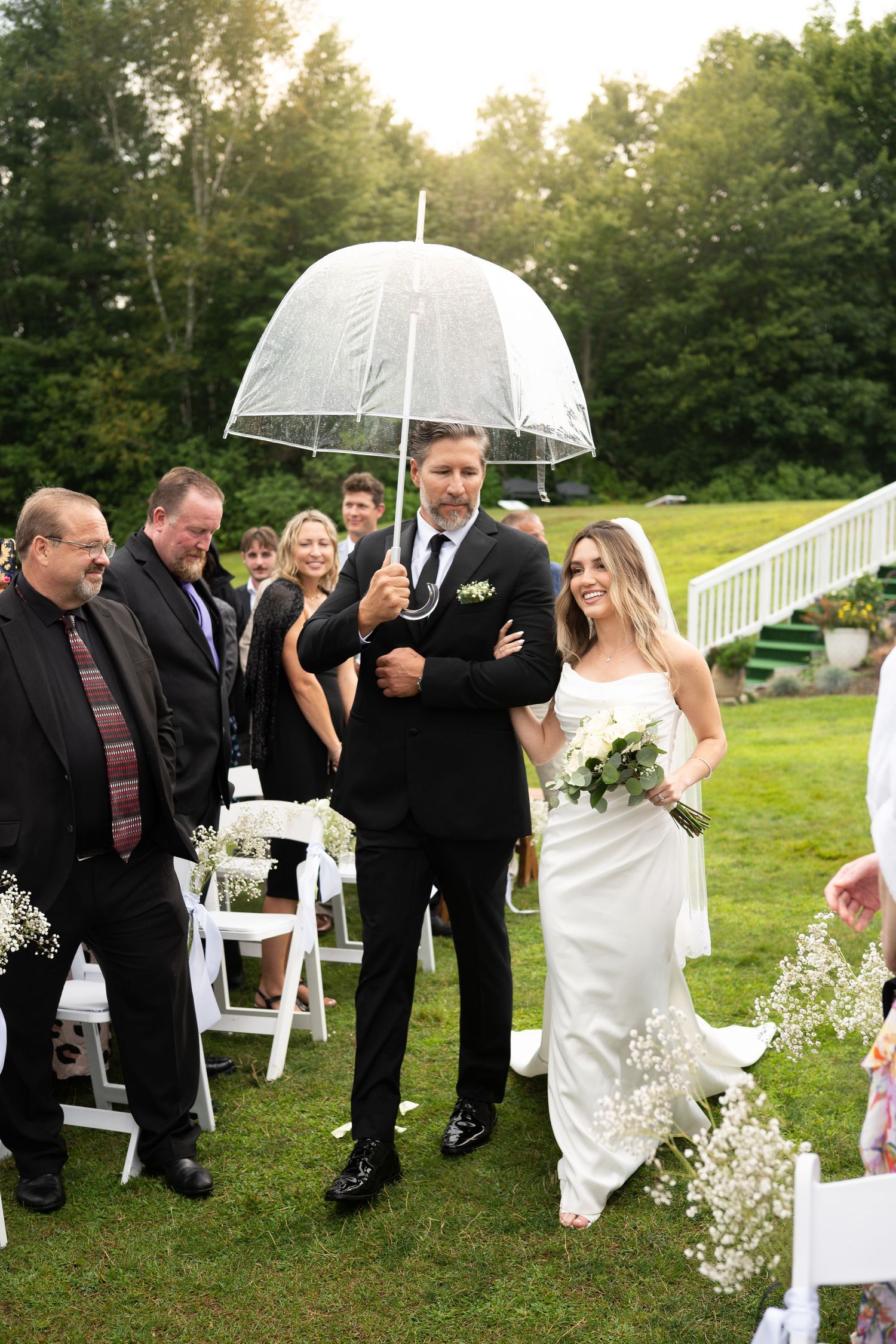 Bride and escort walking down outdoor aisle under clear umbrella on wedding day.