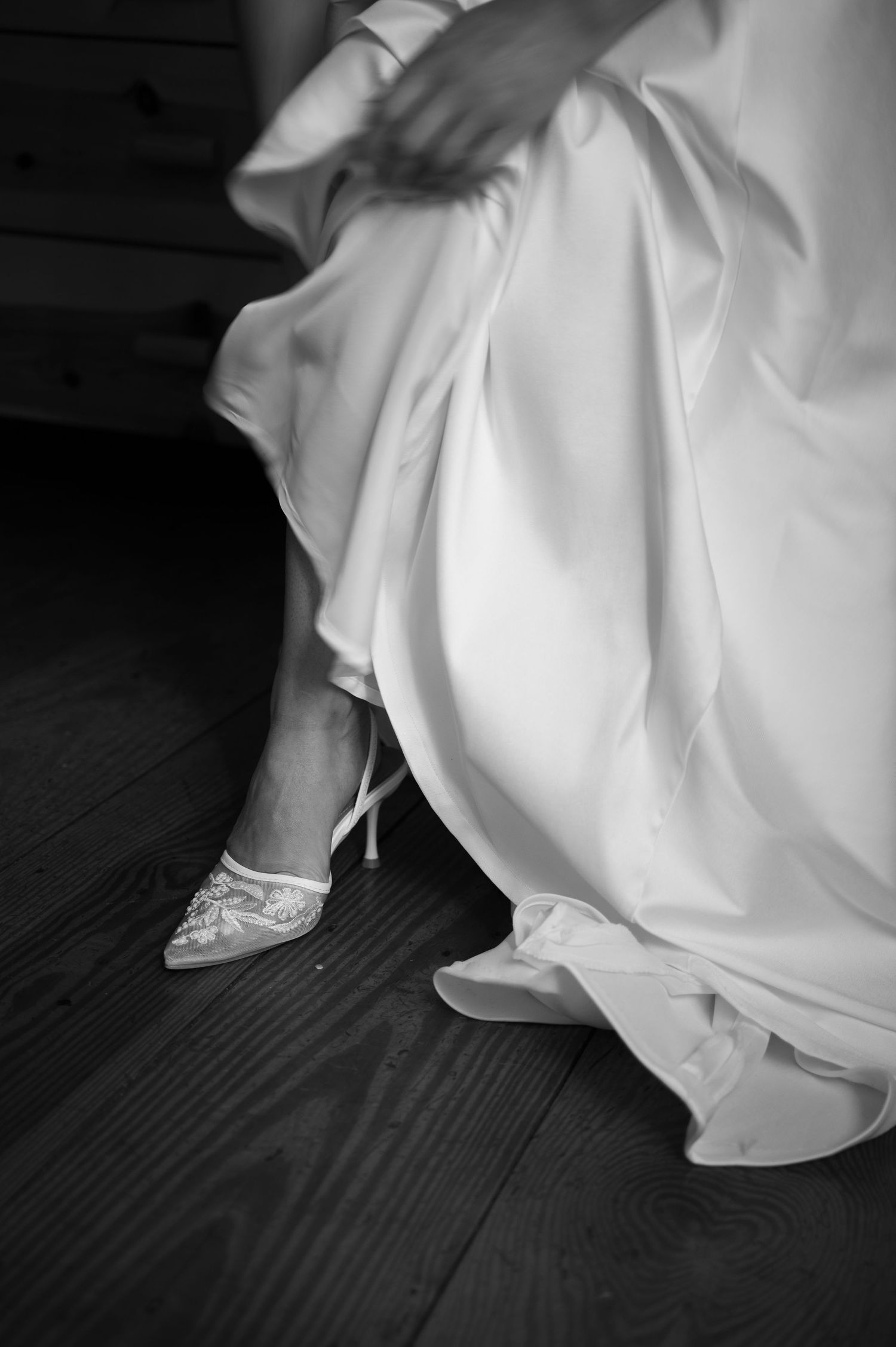 Black and white photo of bride in white gown wearing delicate lace heels on hardwood floor.