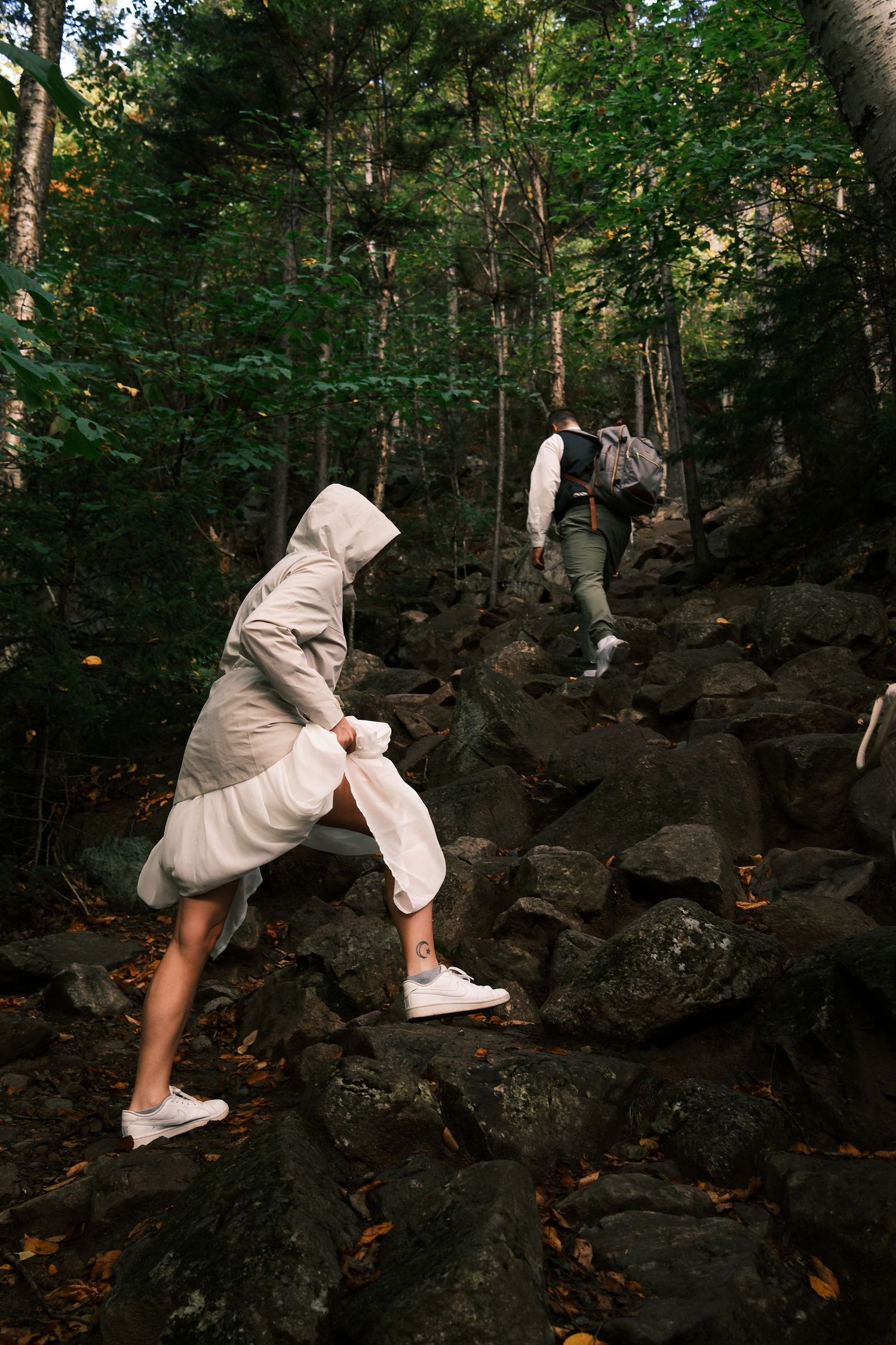 Two hikers climbing rocky forest trail, woman in white hooded coat and sneakers leading the way through lush green woods.