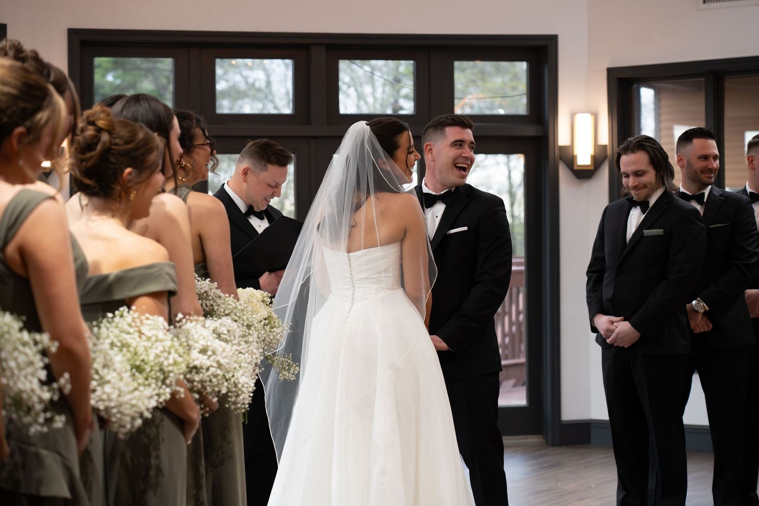 Groom smiles joyfully at bride during wedding ceremony with bridesmaids and groomsmen at altar.