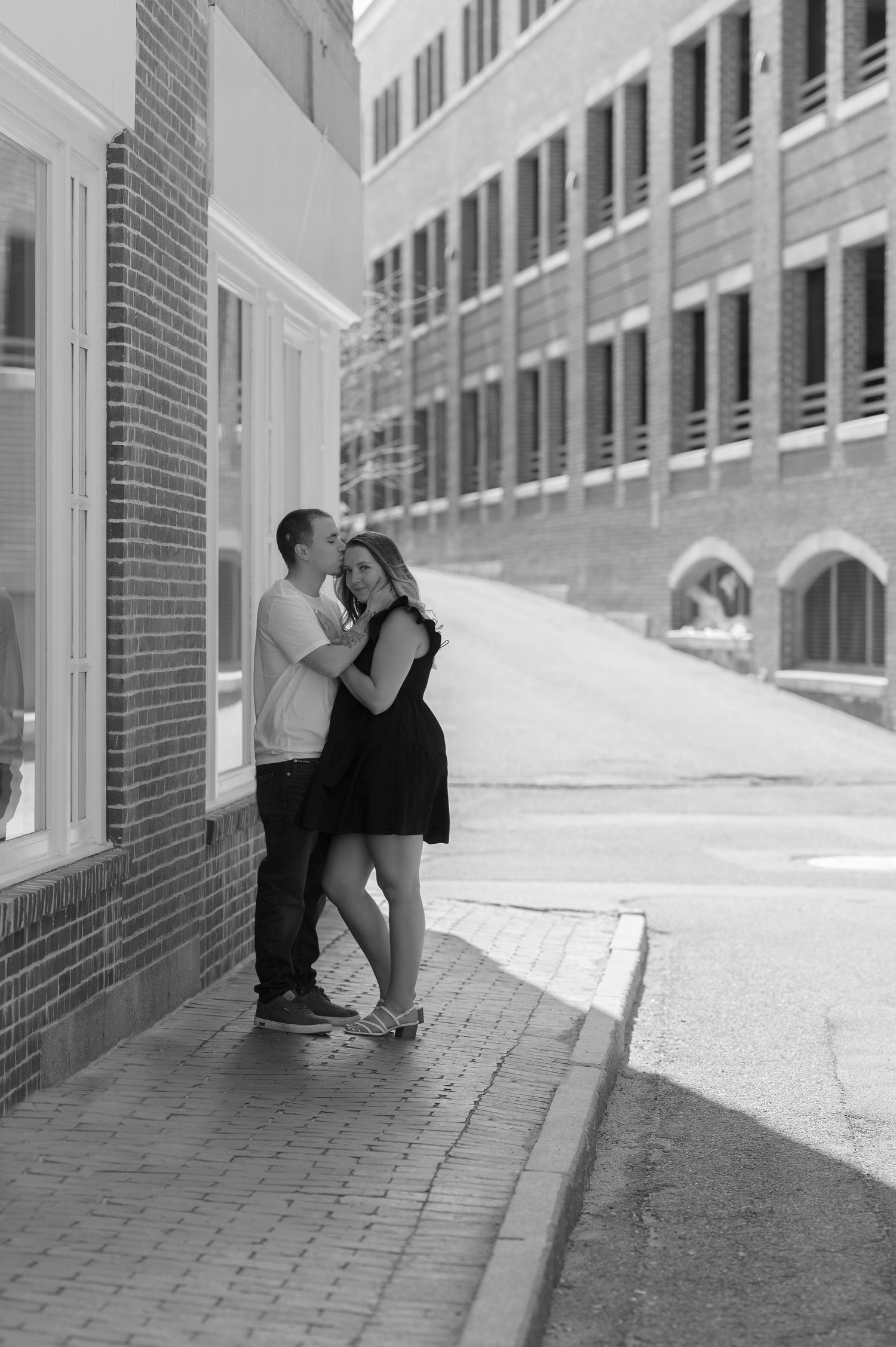 A black and white photo of a couple embracing on a sunlit urban sidewalk near brick buildings.