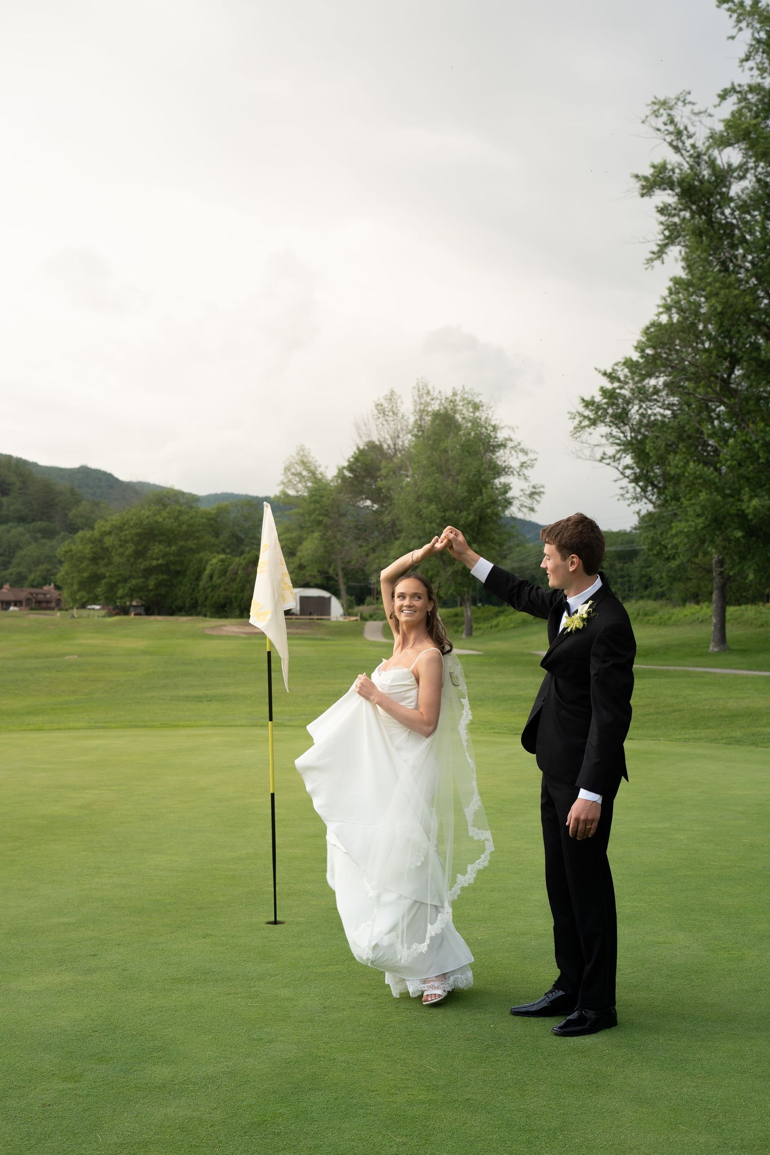 Bride and groom dancing on a lush green golf course, with a white flag nearby and scenic hills in the background.