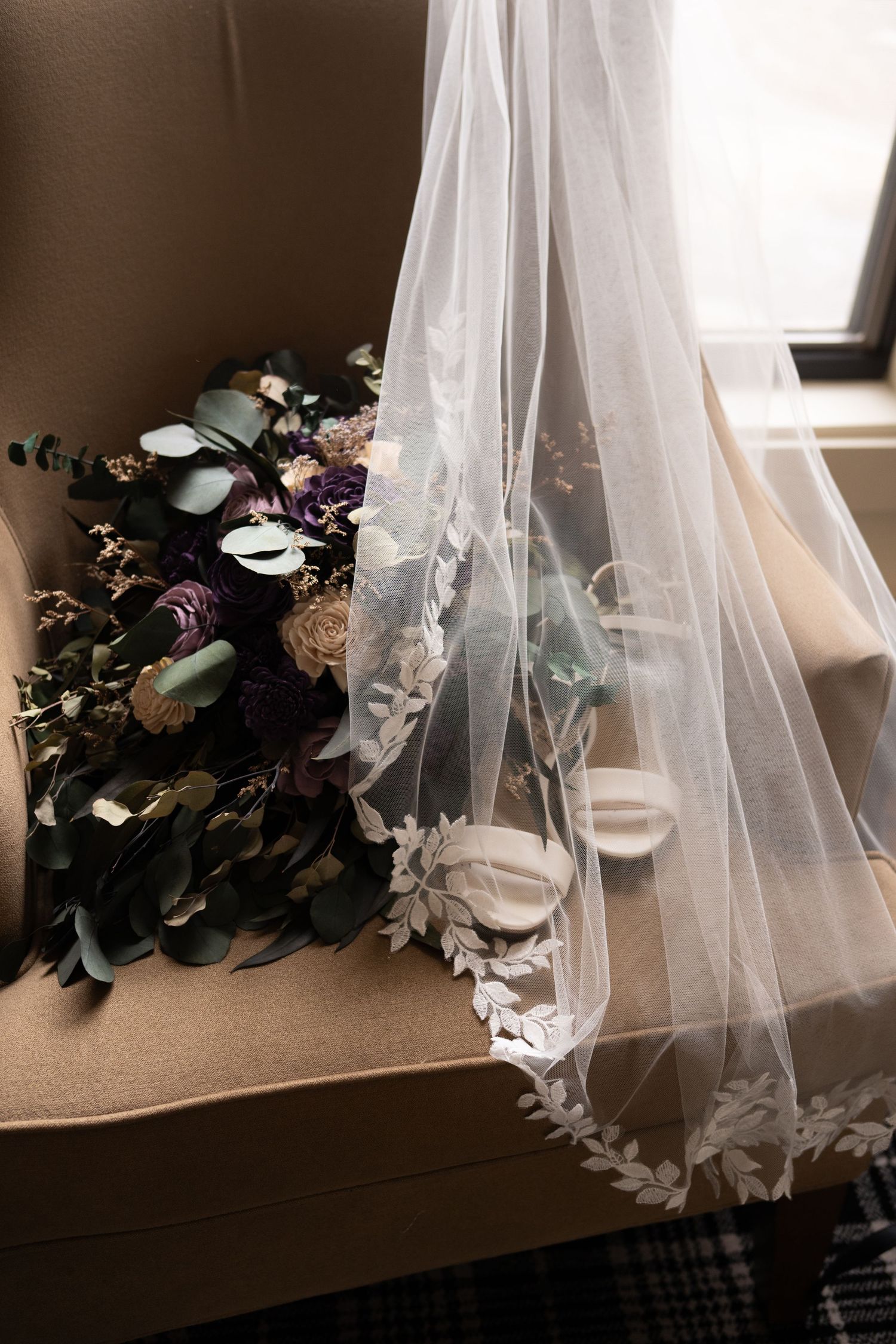 Bridal veil with lace trim draped over chair beside dark floral bouquet with eucalyptus greenery.