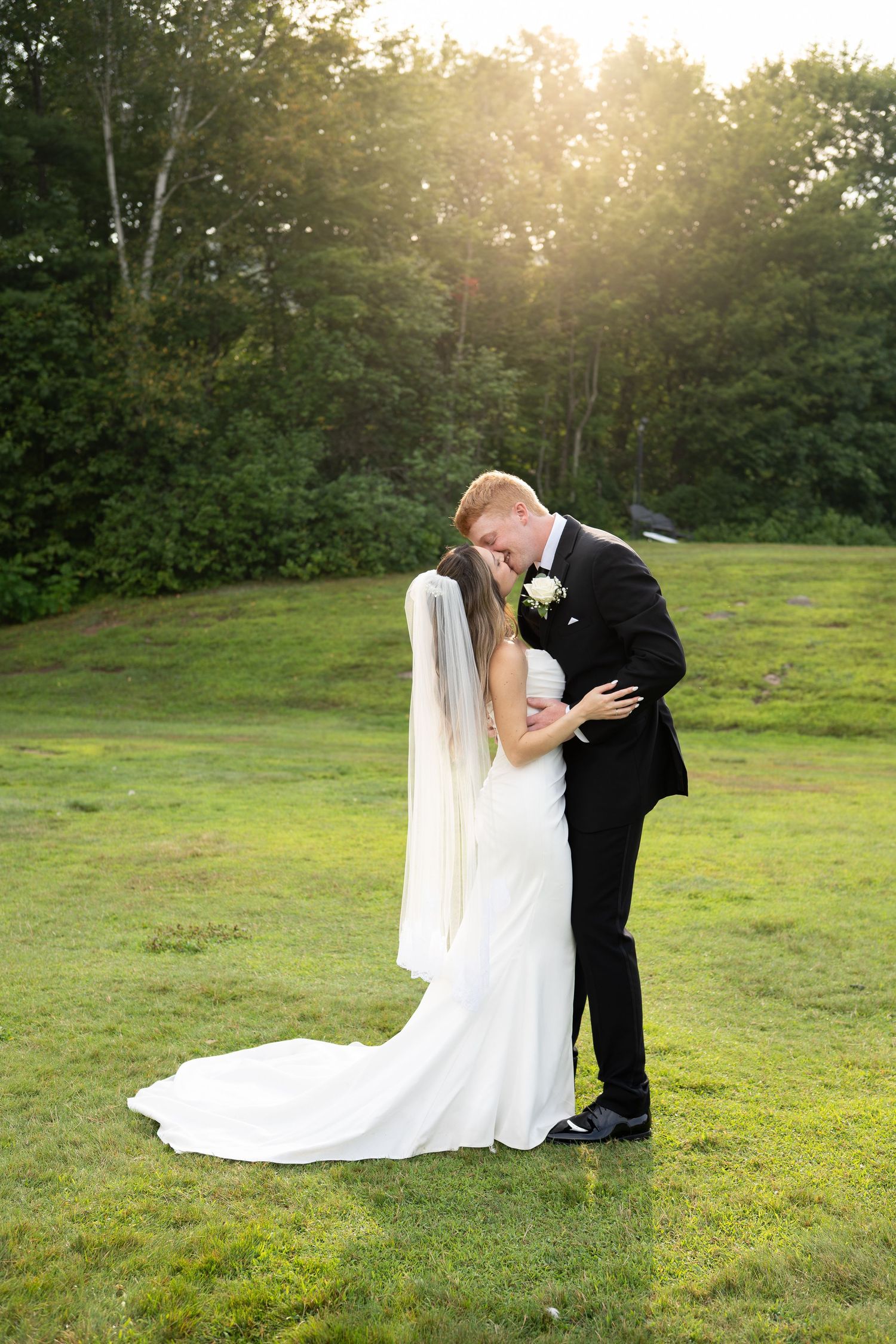 Bride and groom sharing romantic kiss on green lawn with golden sunset backdrop.