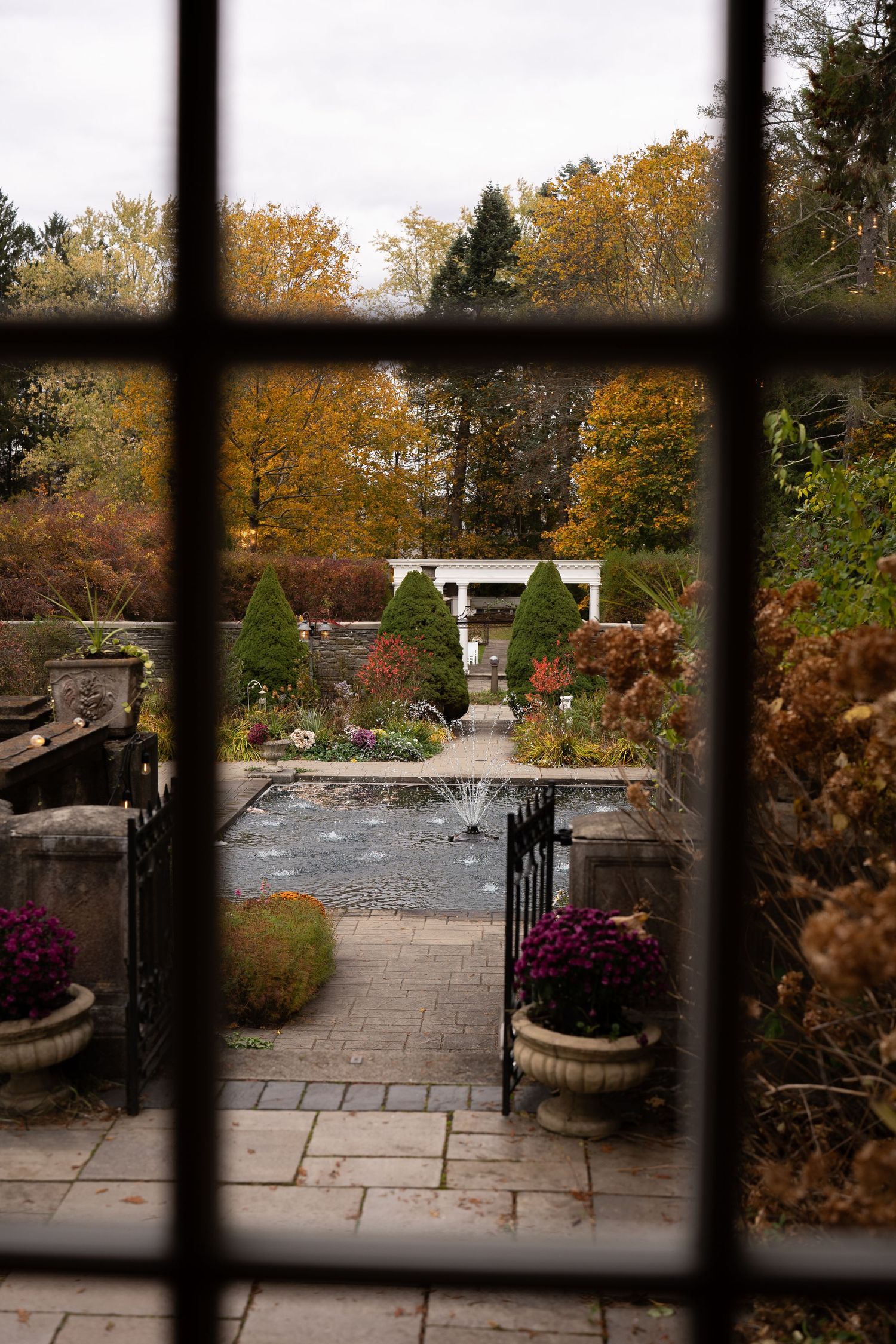 Autumn garden view through iron window frame with fountain, manicured hedges, and colorful fall foliage.