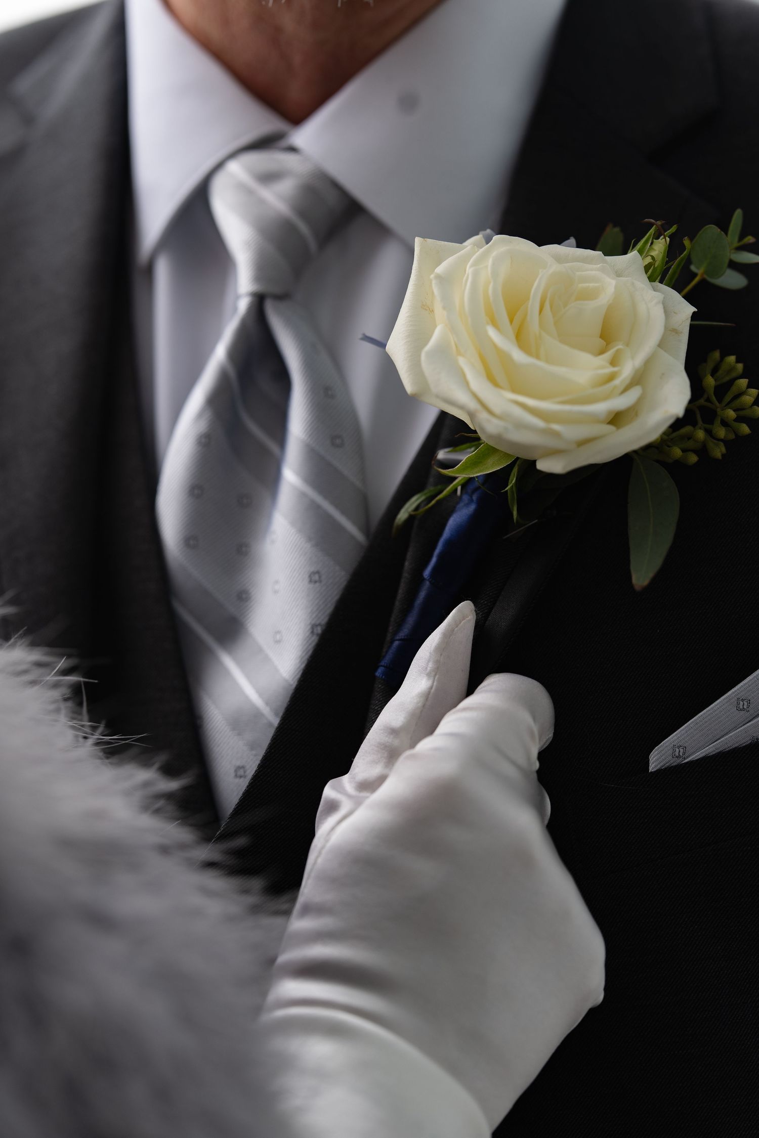 White rose boutonniere being pinned on groom's dark suit lapel by gloved hands.
