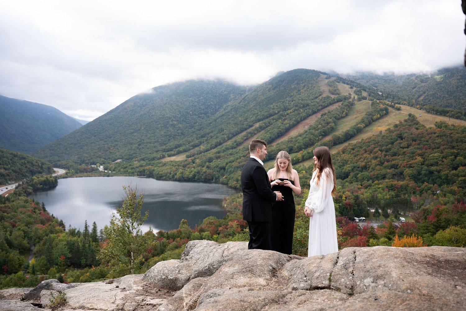 Couple exchanging vows with officiant on rocky mountain overlook above a lake in New Hampshire.