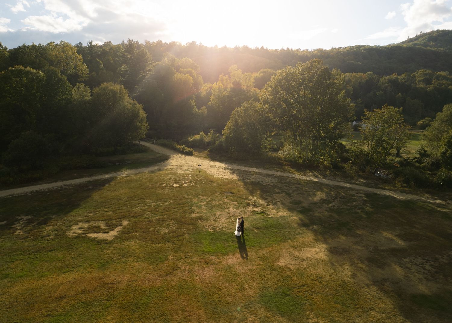 Aerial view of a lone figure standing in a sun-drenched green meadow surrounded by lush forest trees.