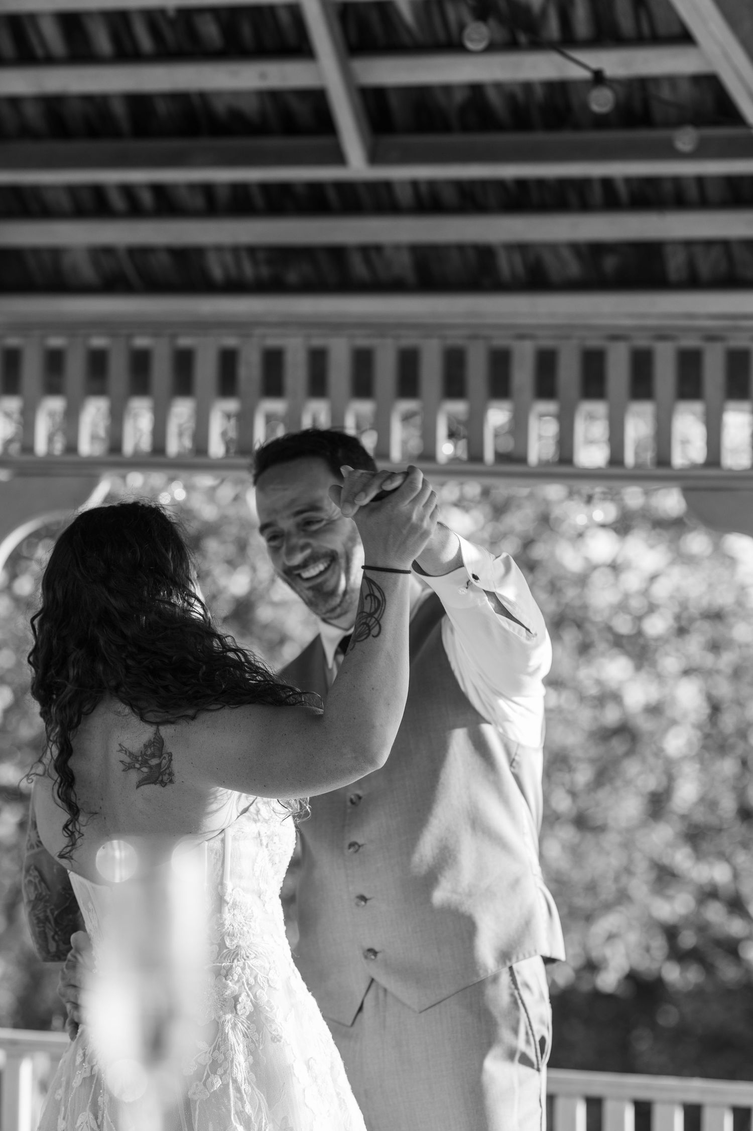 Joyful groom laughing during first dance with bride under rustic pavilion at outdoor wedding reception.