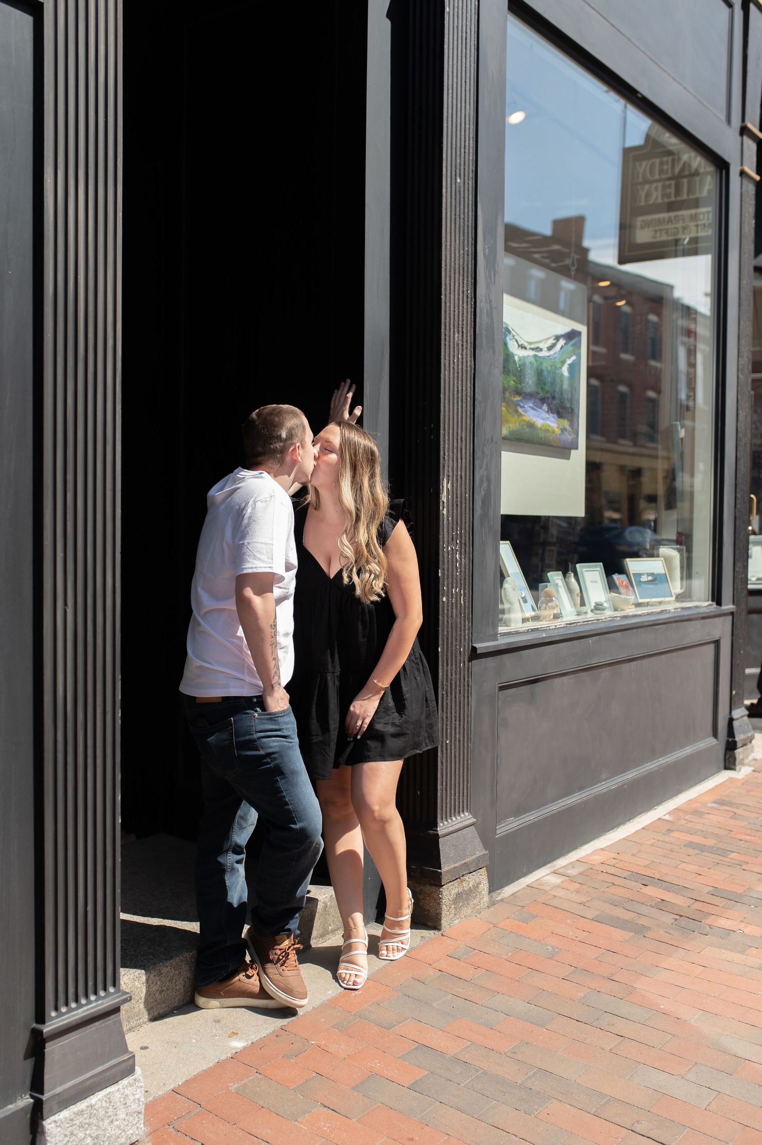 A couple shares a kiss in a storefront doorway on a sunny urban street.