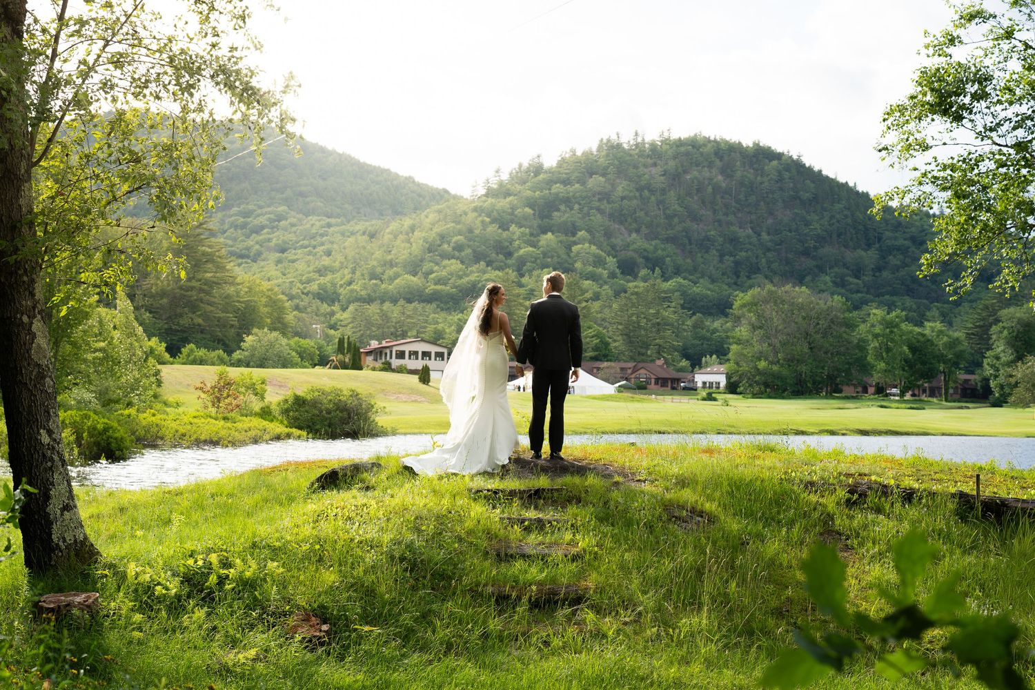 Bride and groom holding hands overlooking a lush green valley with a river and mountain backdrop.