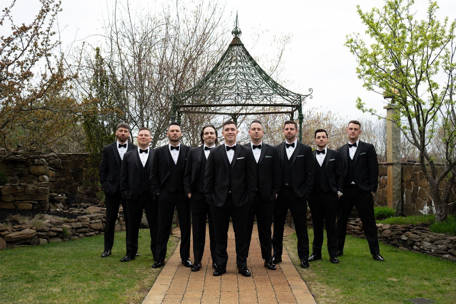 Groomsmen in matching black tuxedos and bow ties pose together in front of garden gazebo.