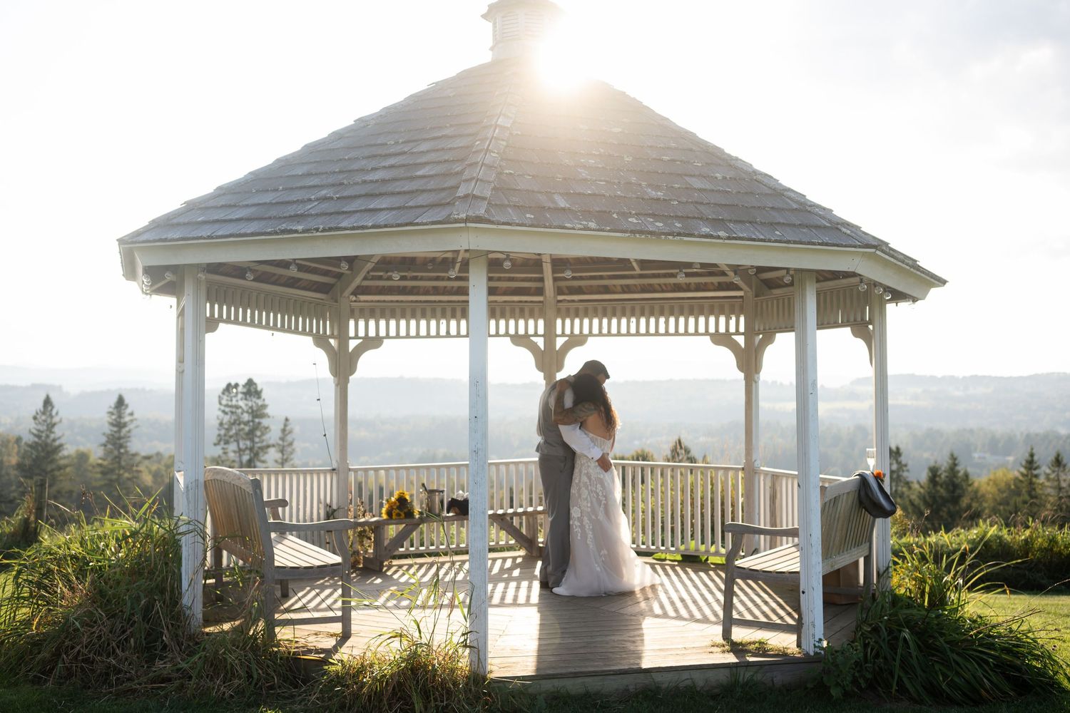 Bride and groom embracing inside a white gazebo with panoramic countryside views.