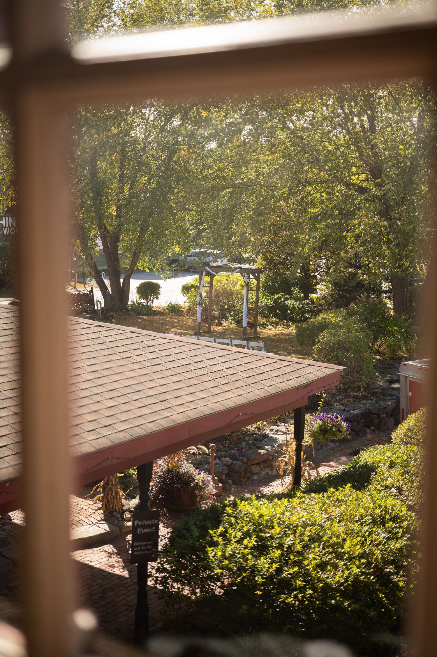 Sunlit garden venue viewed through a window frame, featuring a white gazebo surrounded by lush green trees.